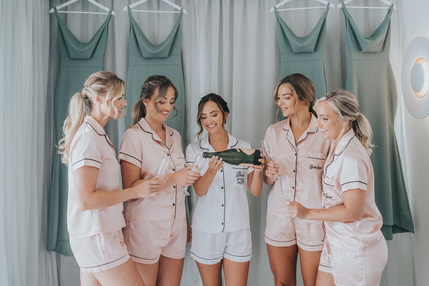 Group wearing matching pink pajama sets getting ready in a room with bridesmaid dresses.