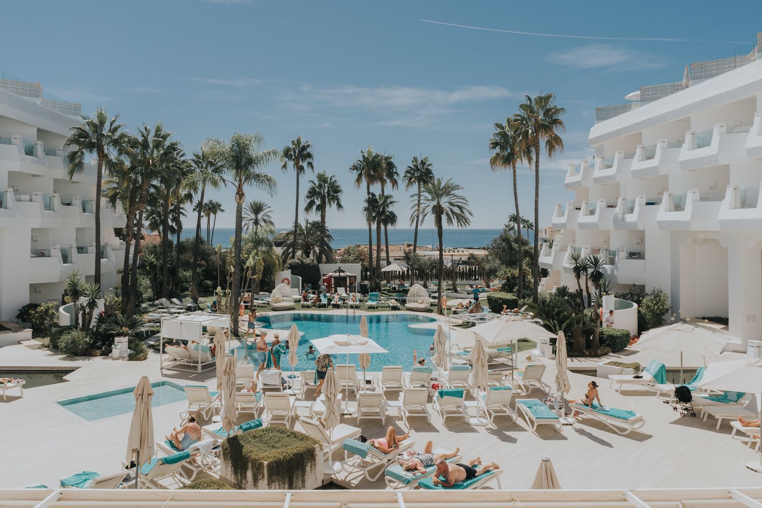 Luxurious resort pool area surrounded by white buildings and palm trees under blue sky.