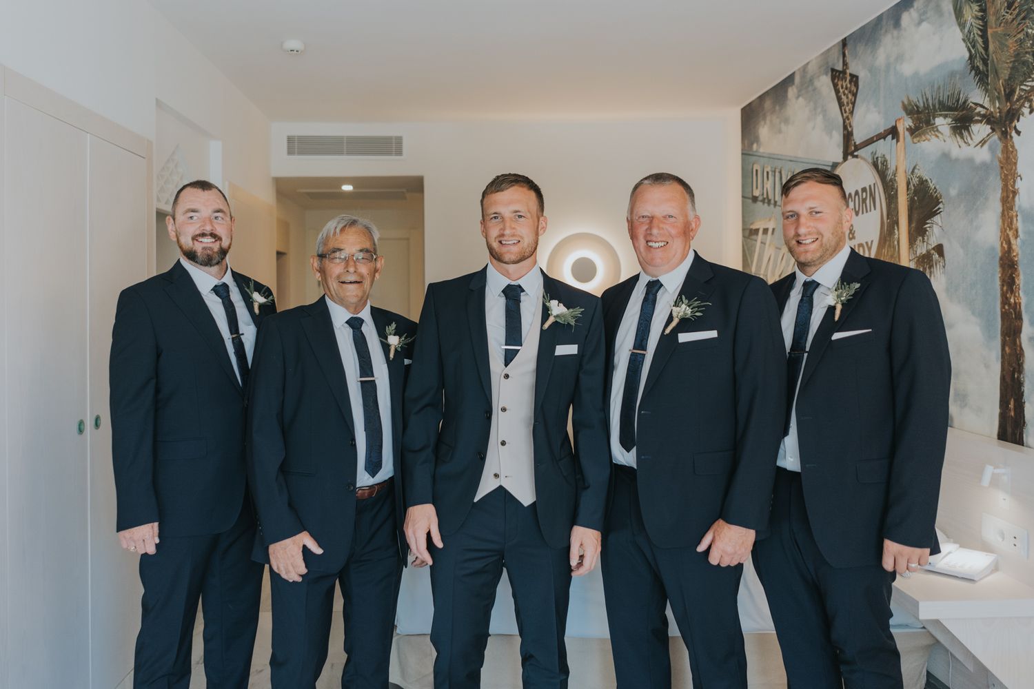 Groomsmen in formal suits and ties pose together in elegant indoor setting.