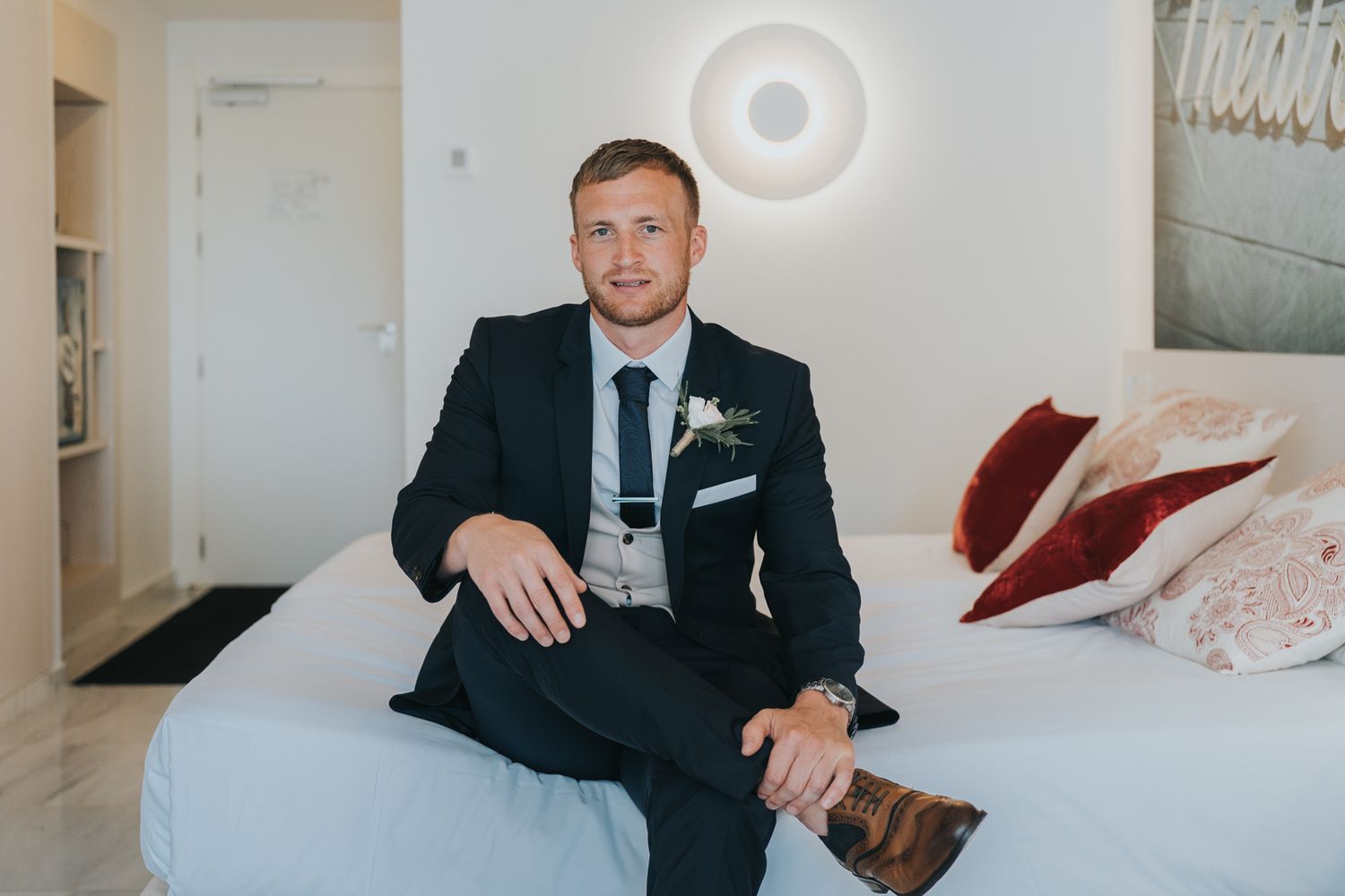 Person in dark suit and tie sits on white bed in modern bedroom.