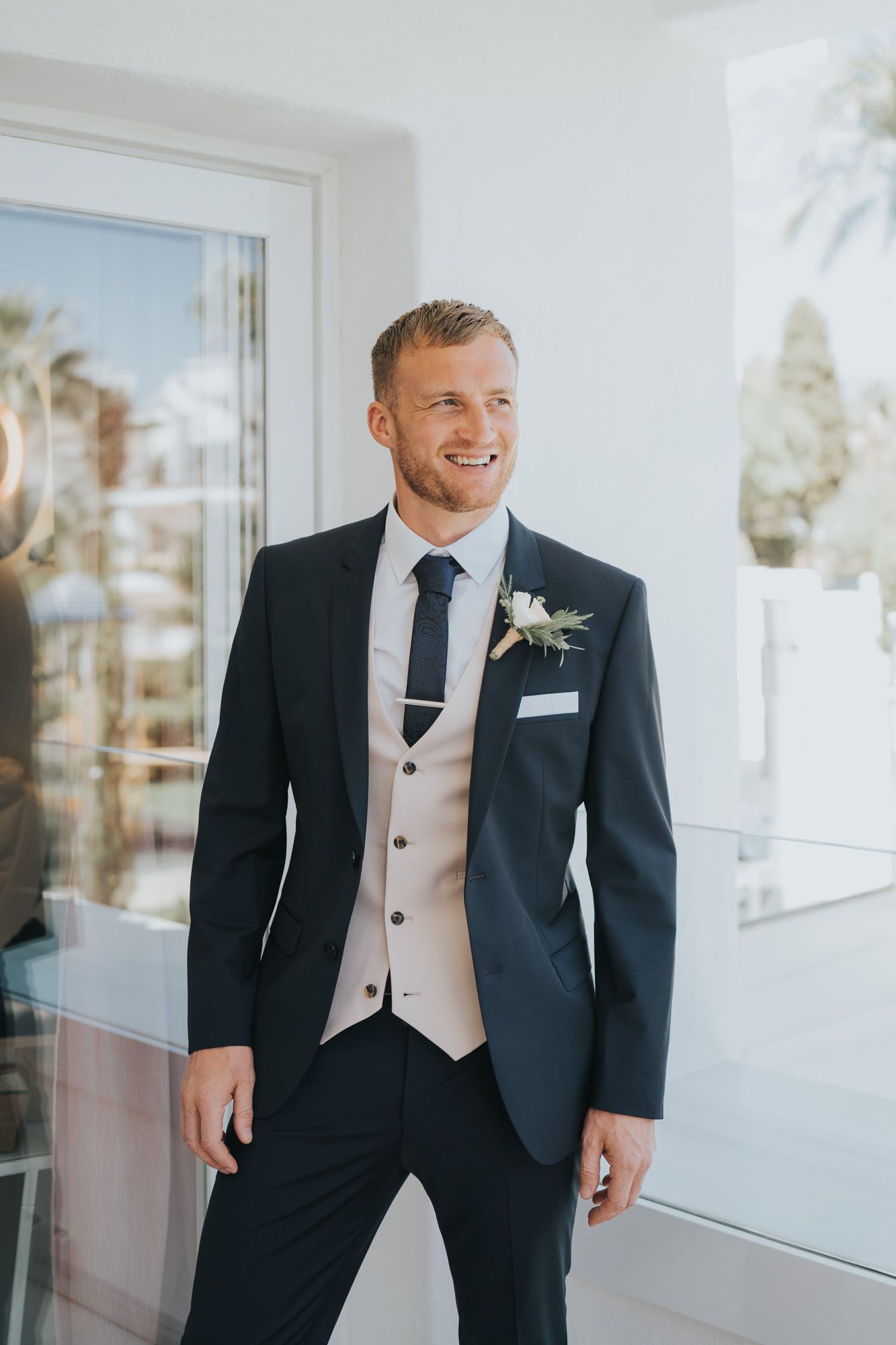 Groom in navy suit with light vest poses near window on wedding day.