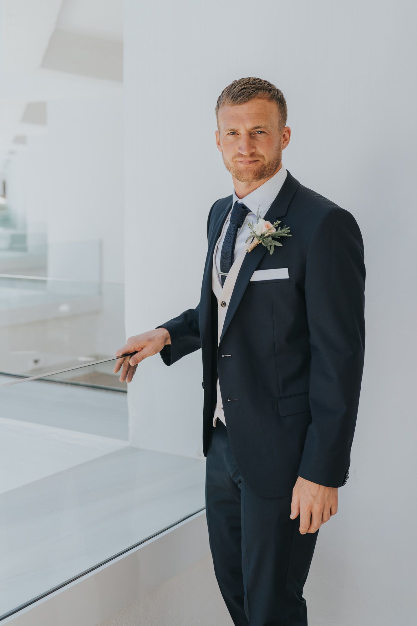 A groom in a classic navy suit and grey waistcoat stands confidently in a modern white interior setting.