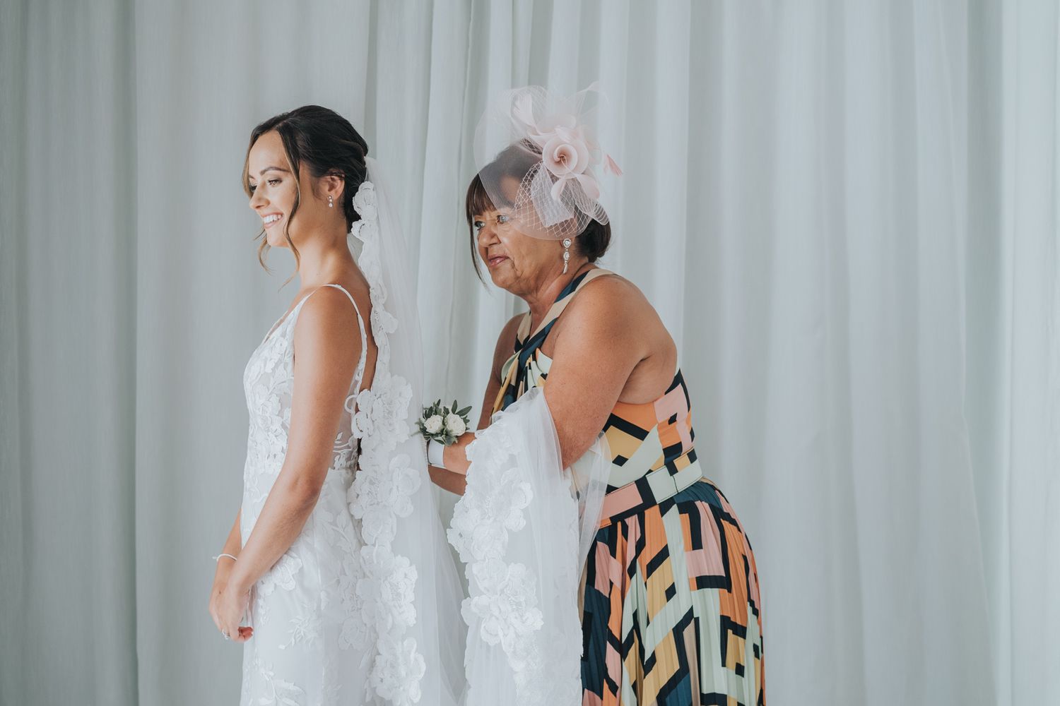 Two people stand together as one helps adjust a white wedding dress near white curtains.