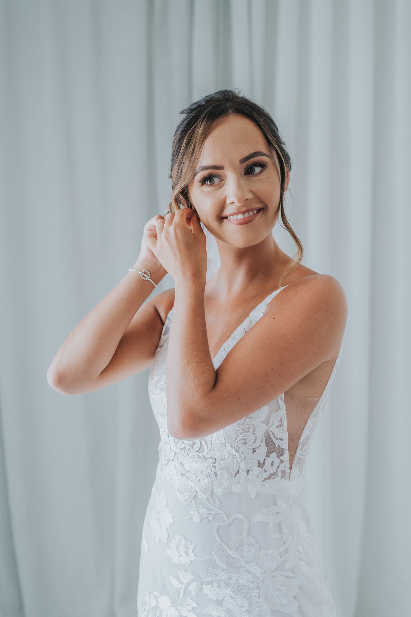 Elegant bride in lace wedding dress putting on earrings against white backdrop.