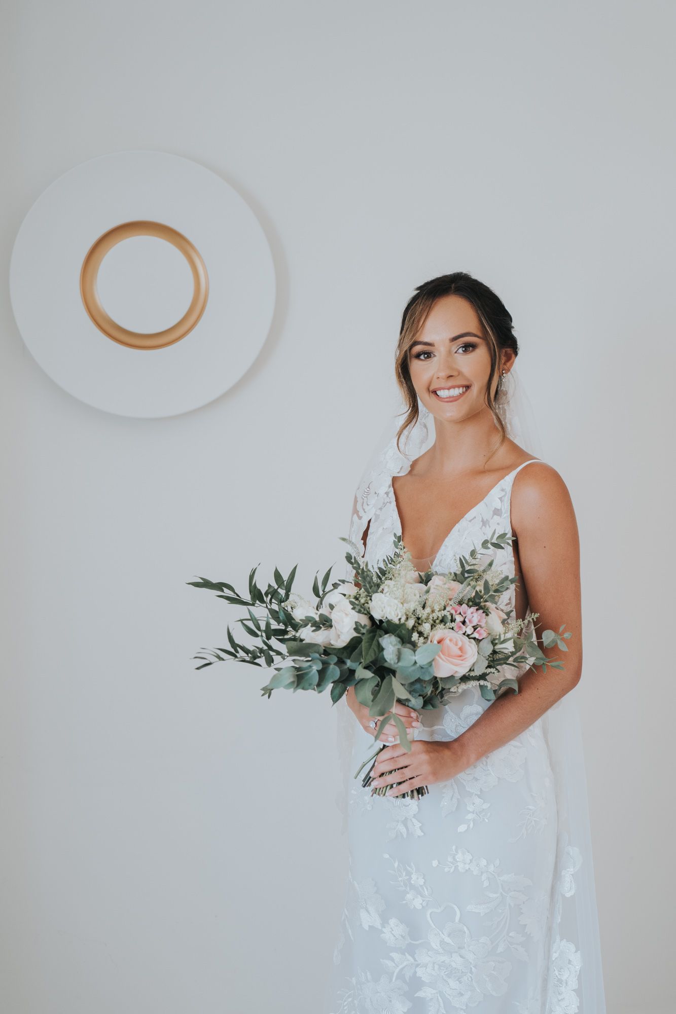 Bride holds elegant white and pink bouquet against minimalist white wall with circular art piece.