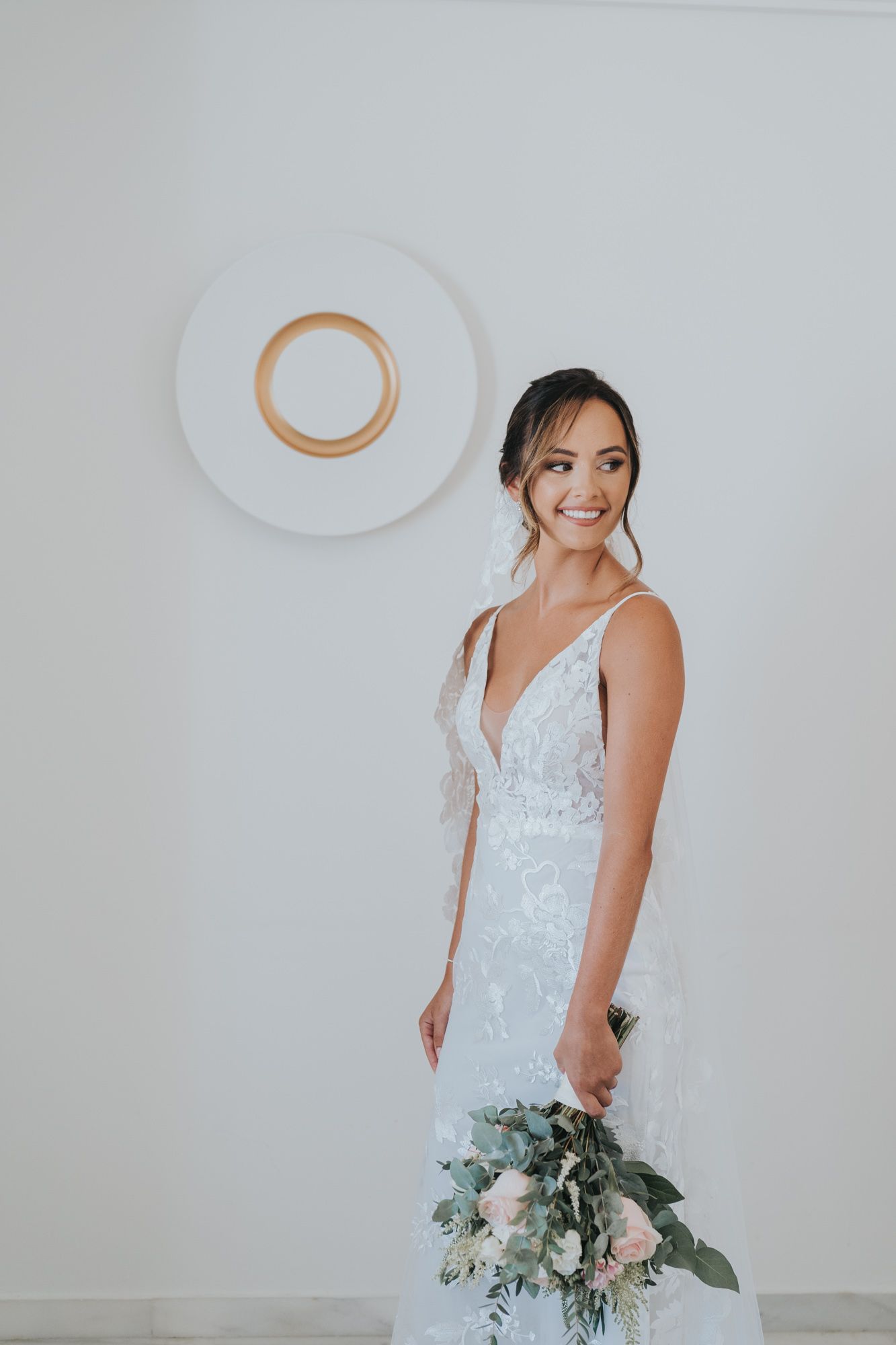 A person in a white lace wedding dress poses with a bouquet against a minimalist backdrop.