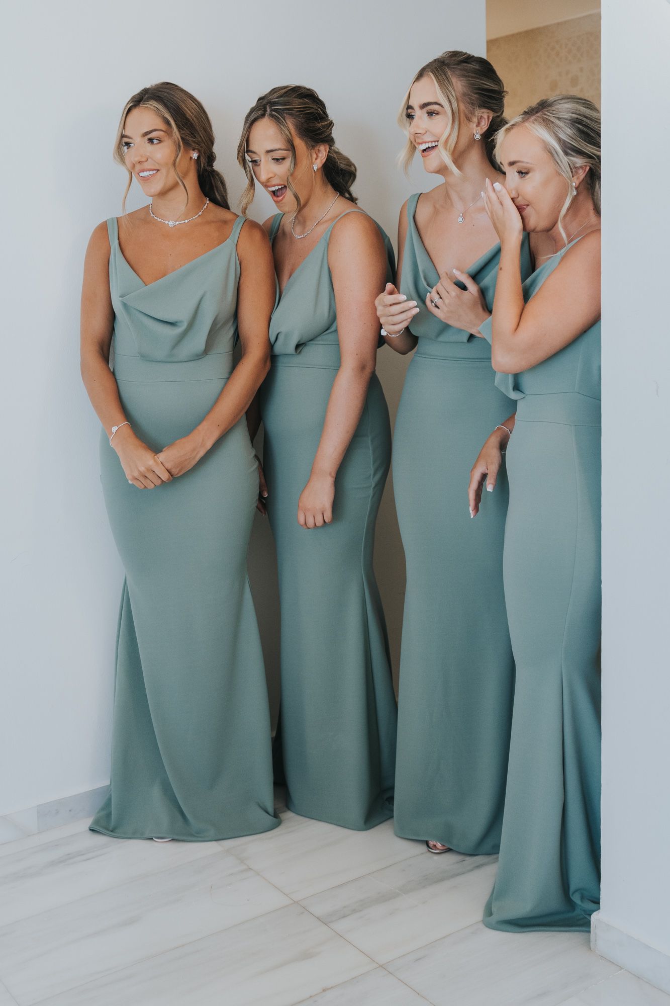 Four bridesmaids in matching sage green floor-length dresses stand together laughing and sharing a candid moment.