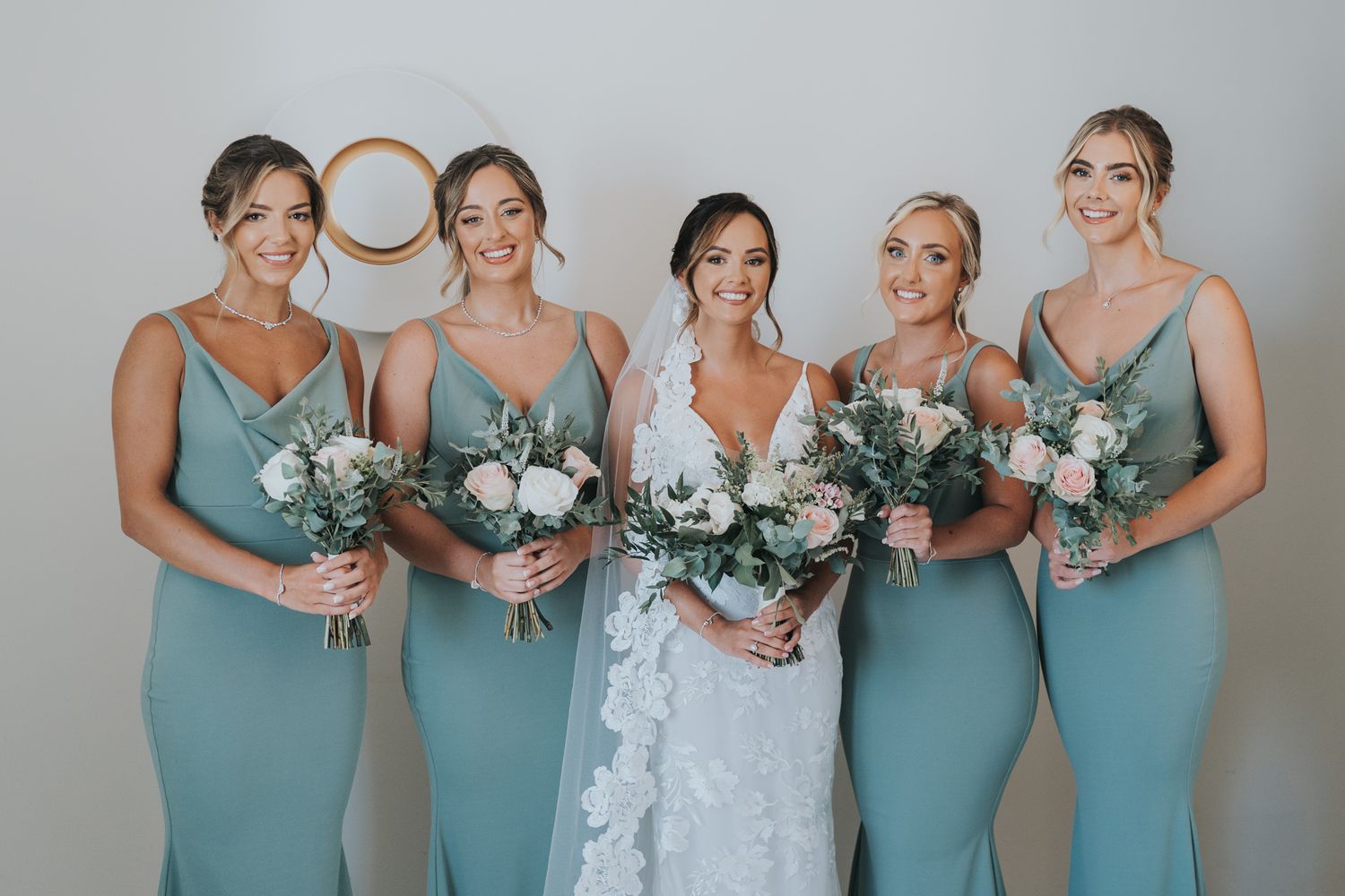 Wedding party in matching seafoam green dresses holding elegant white and green floral bouquets pose for formal photo.