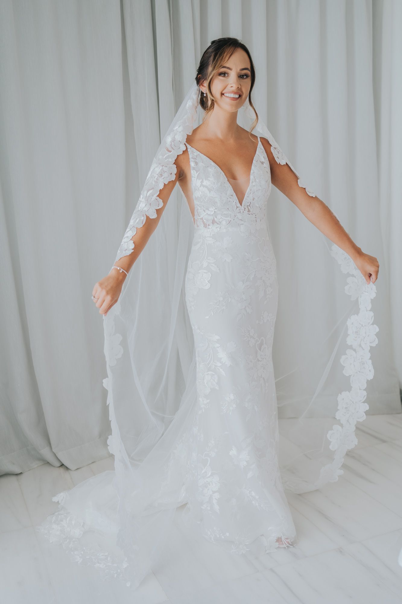Elegant lace wedding gown with deep V-neckline and flowing veil against white backdrop.