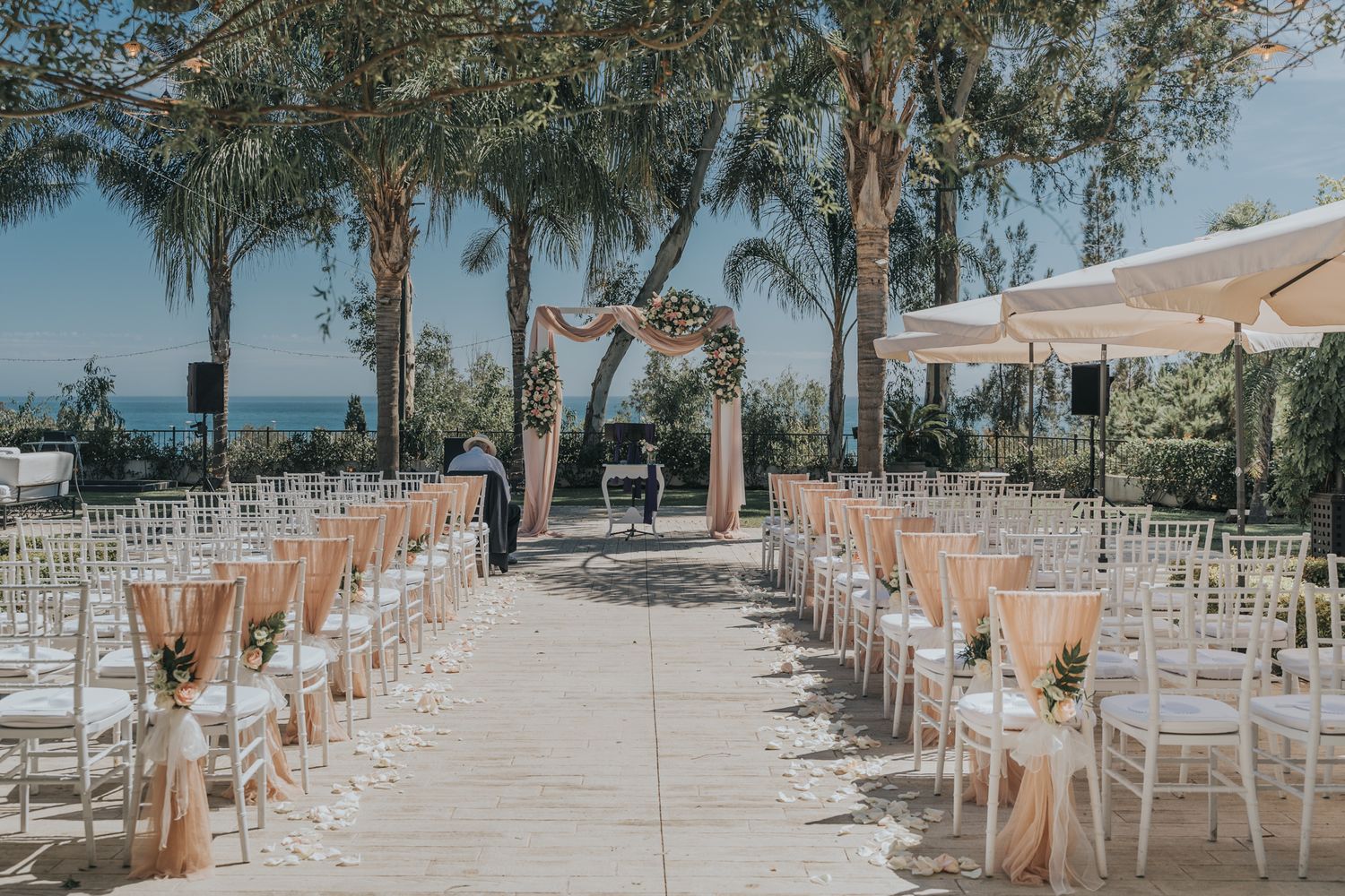 An elegant outdoor wedding ceremony setup with white chairs on a stone patio overlooking the ocean through palm trees.