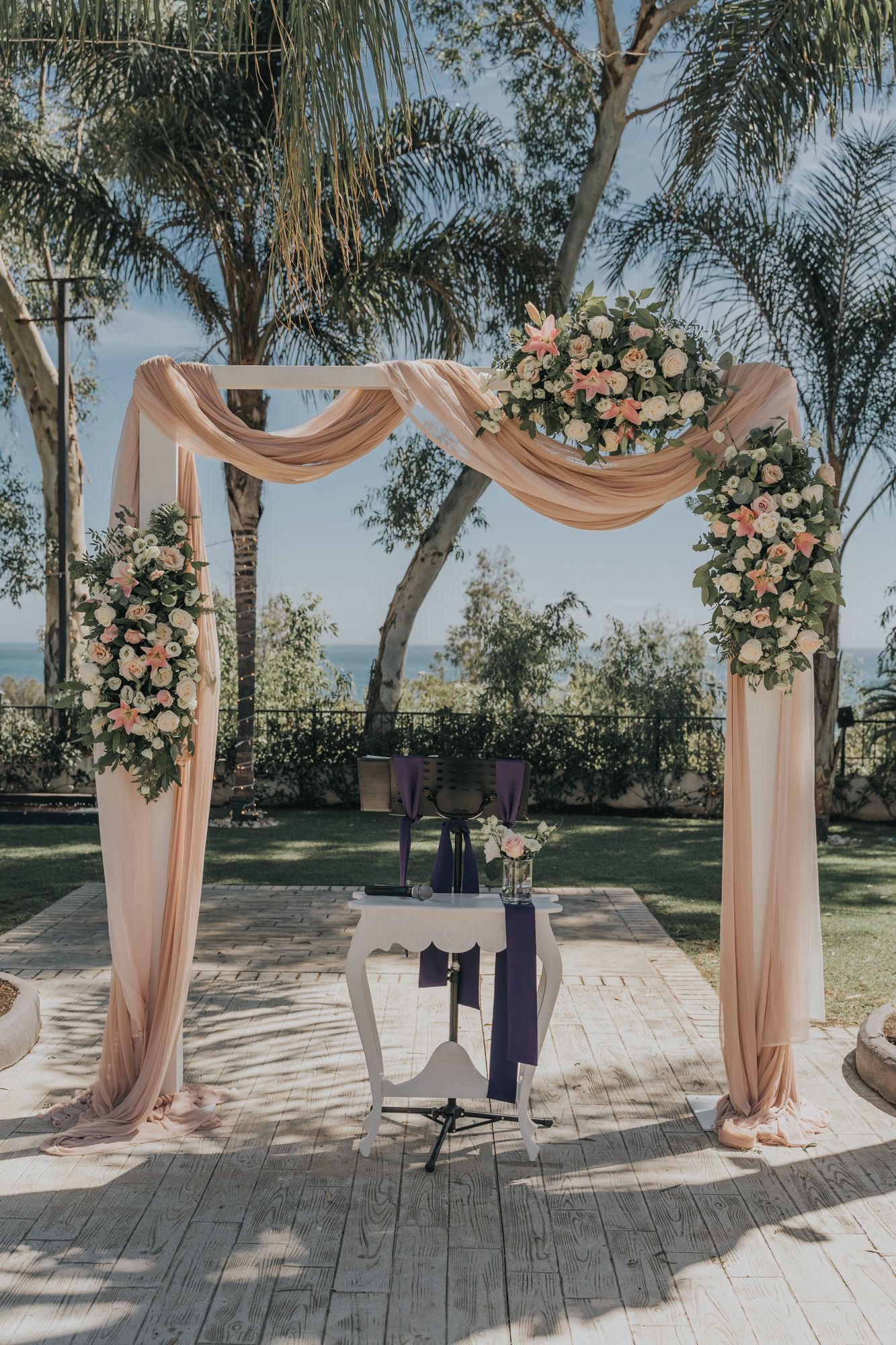 Romantic wedding arch with blush draping and floral arrangements against palm tree backdrop.
