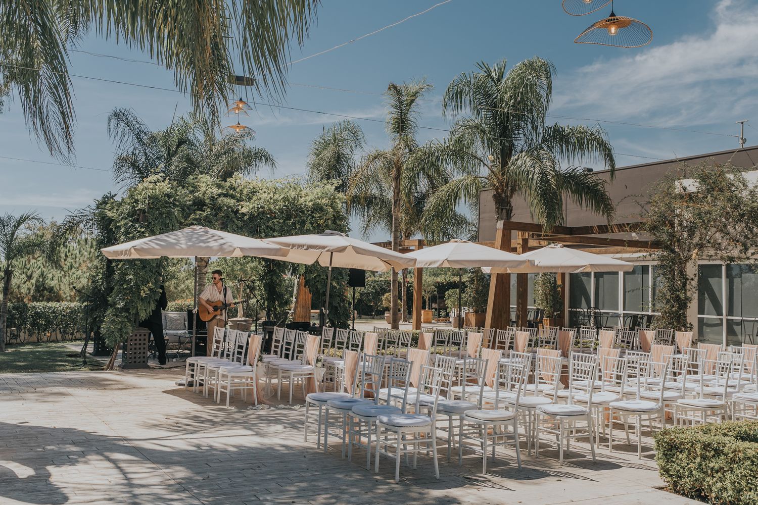Outdoor wedding ceremony setup with white chairs and umbrellas under palm trees.
