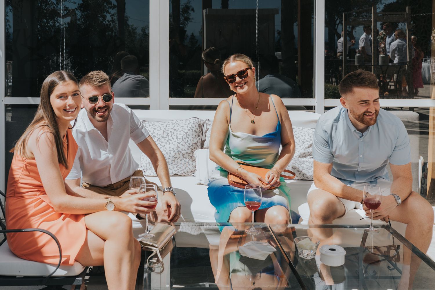 A group of friends sitting outdoors on a patio sofa enjoying drinks together on a sunny day.