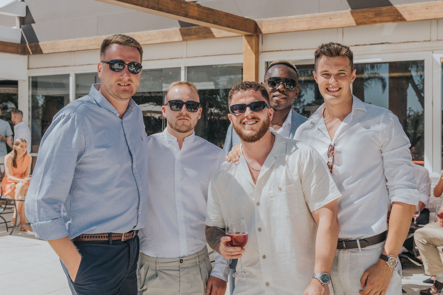 Group of friends in casual attire enjoying drinks at an outdoor venue wearing sunglasses.
