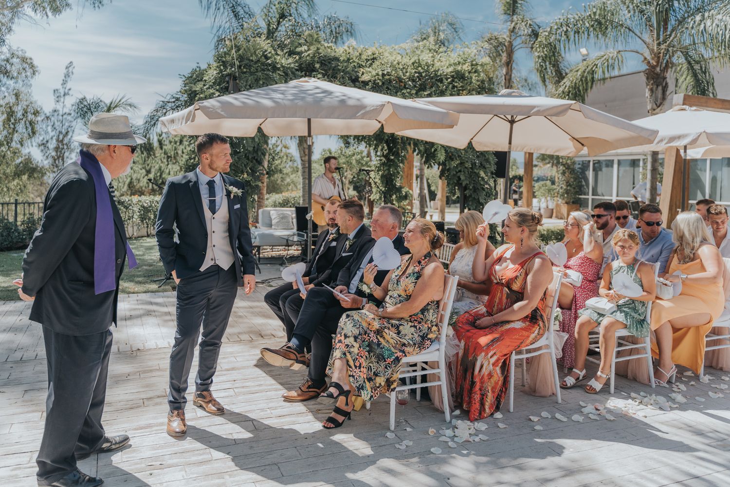 Outdoor wedding ceremony with guests seated under white umbrellas on a sunny patio.