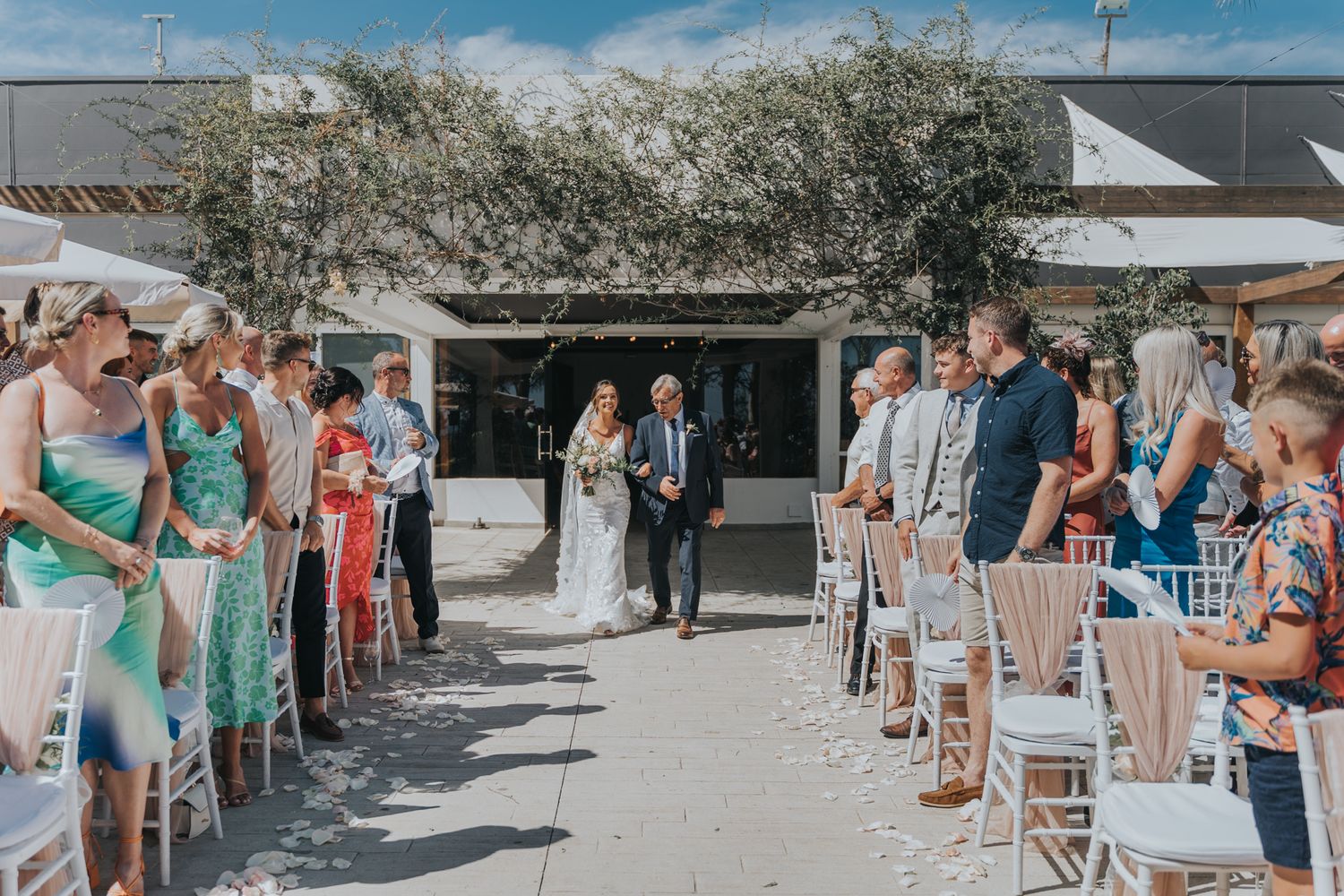 Wedding ceremony aisle lined with guests in casual attire under a rustic outdoor arch.