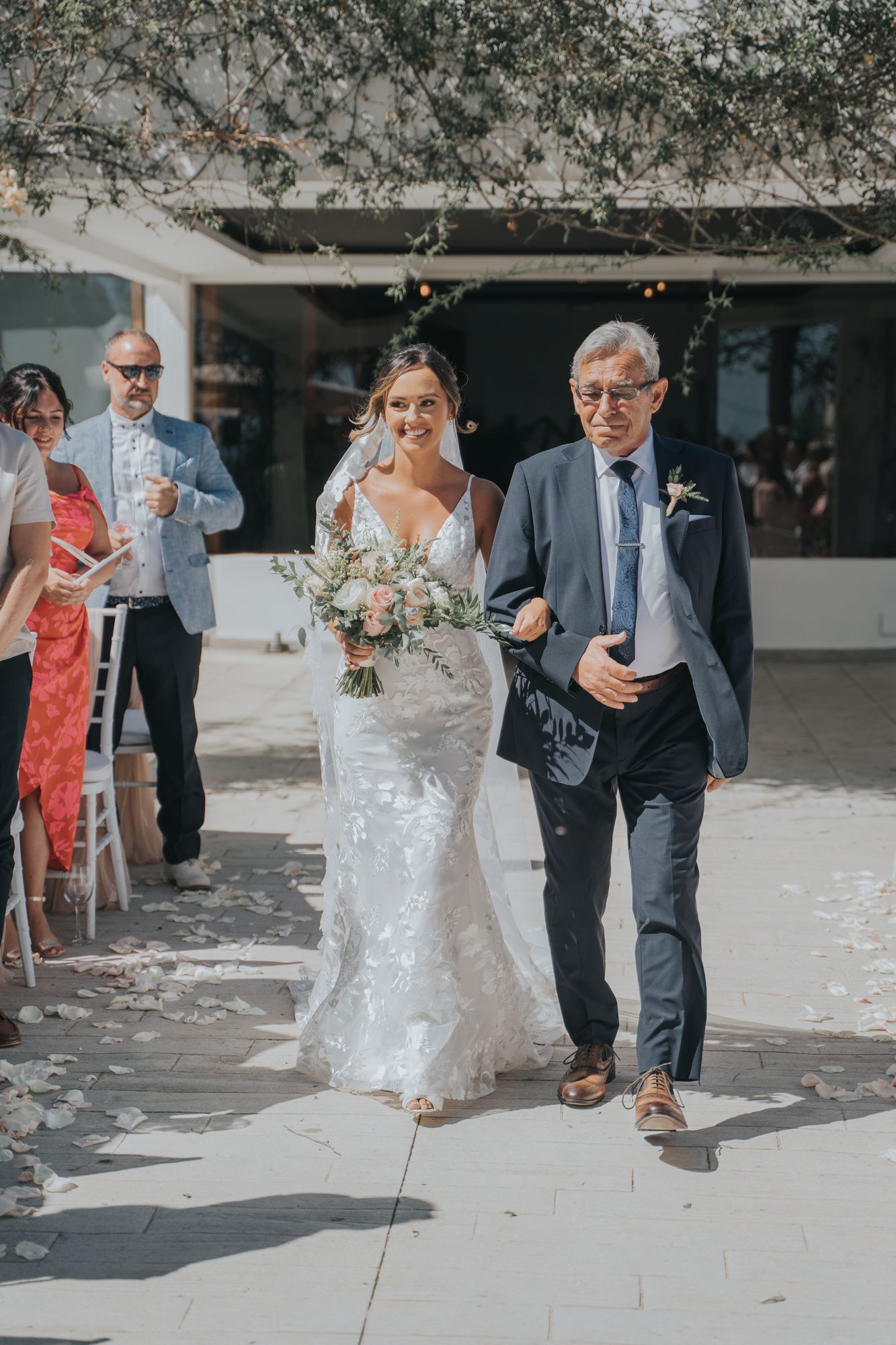 A bride in a lace dress walks down a sunlit path carrying a white bouquet while escorted by a gentleman.