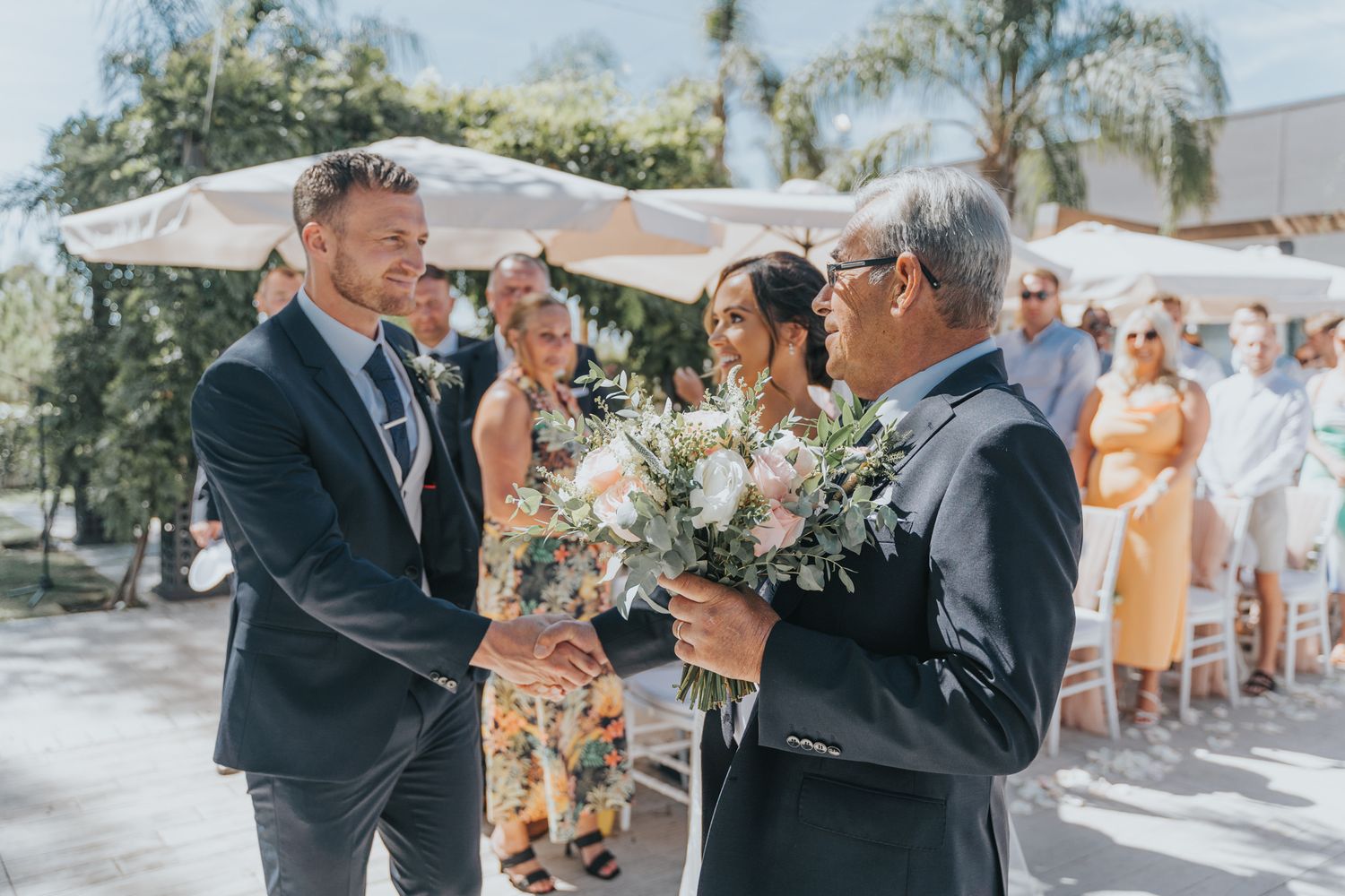 Groomsmen exchange handshakes and bouquets at an outdoor wedding ceremony under white canopies.