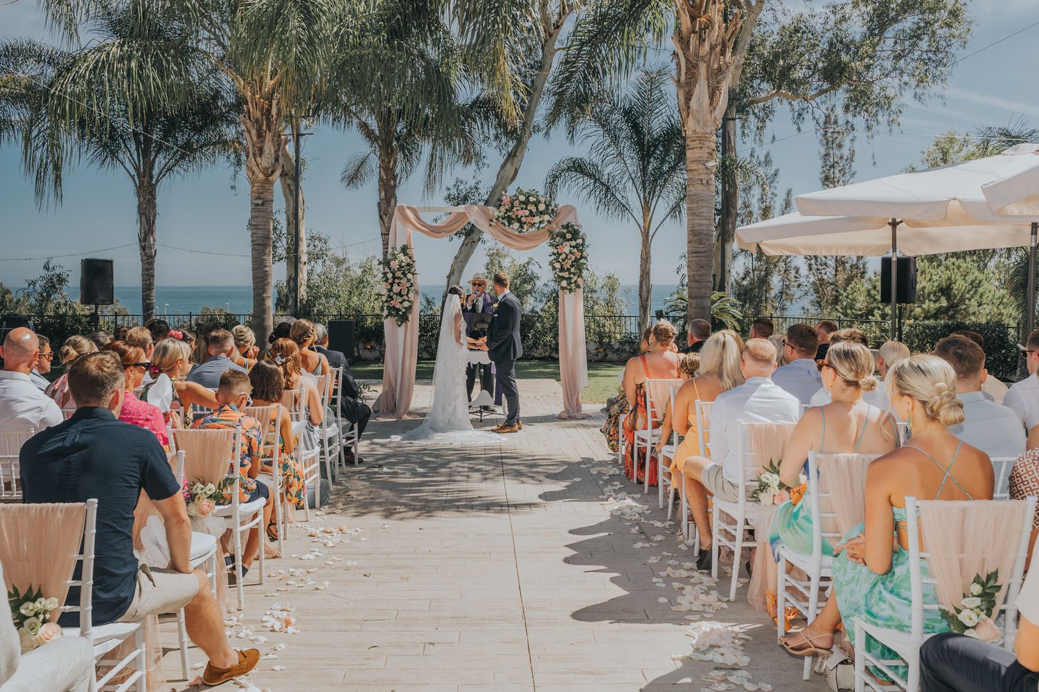 Palm trees line an outdoor wedding ceremony with white chairs and a floral arch at the altar.