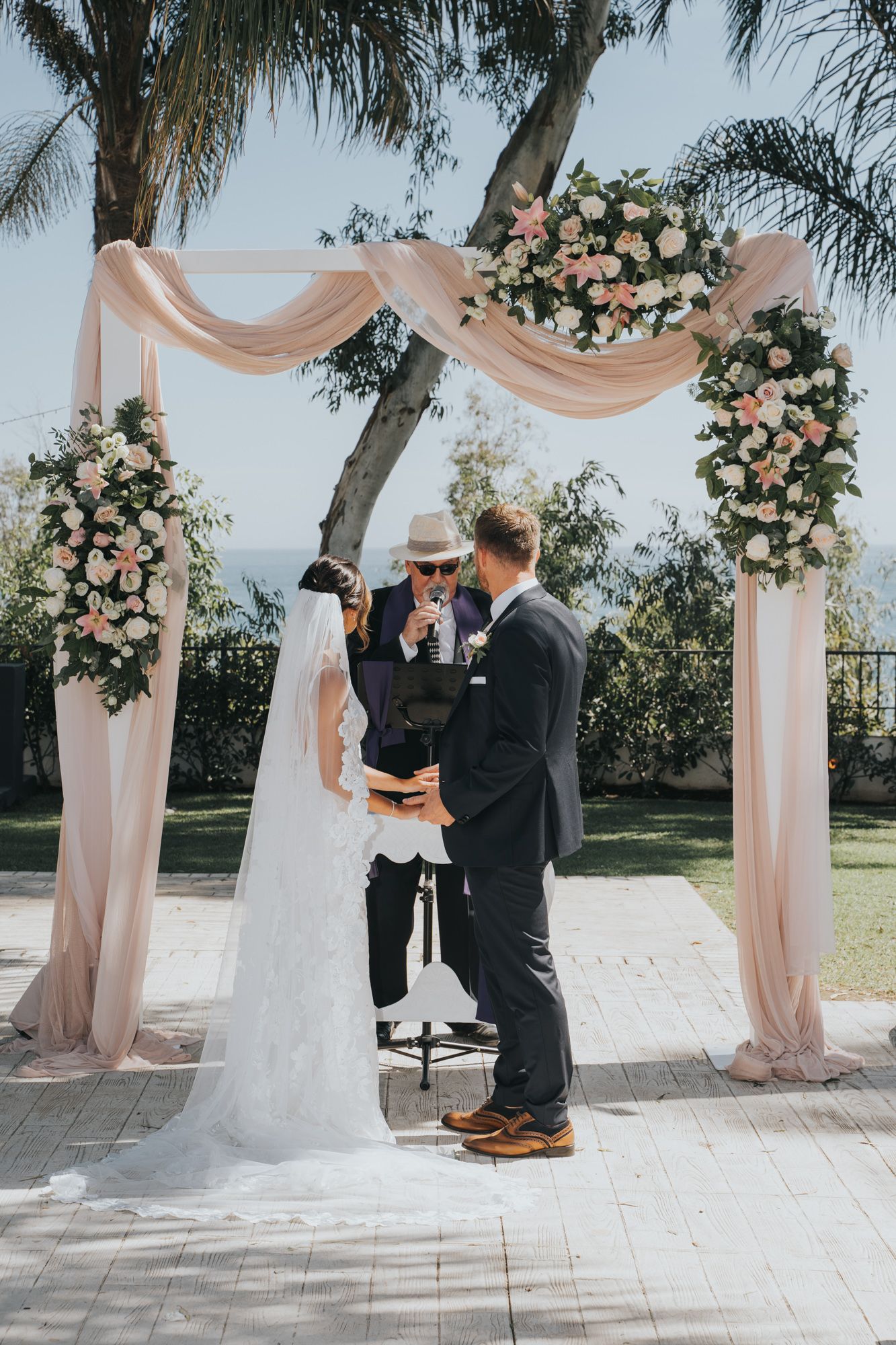 Wedding ceremony under a blush pink draped arch decorated with pink and white roses in an outdoor garden setting.