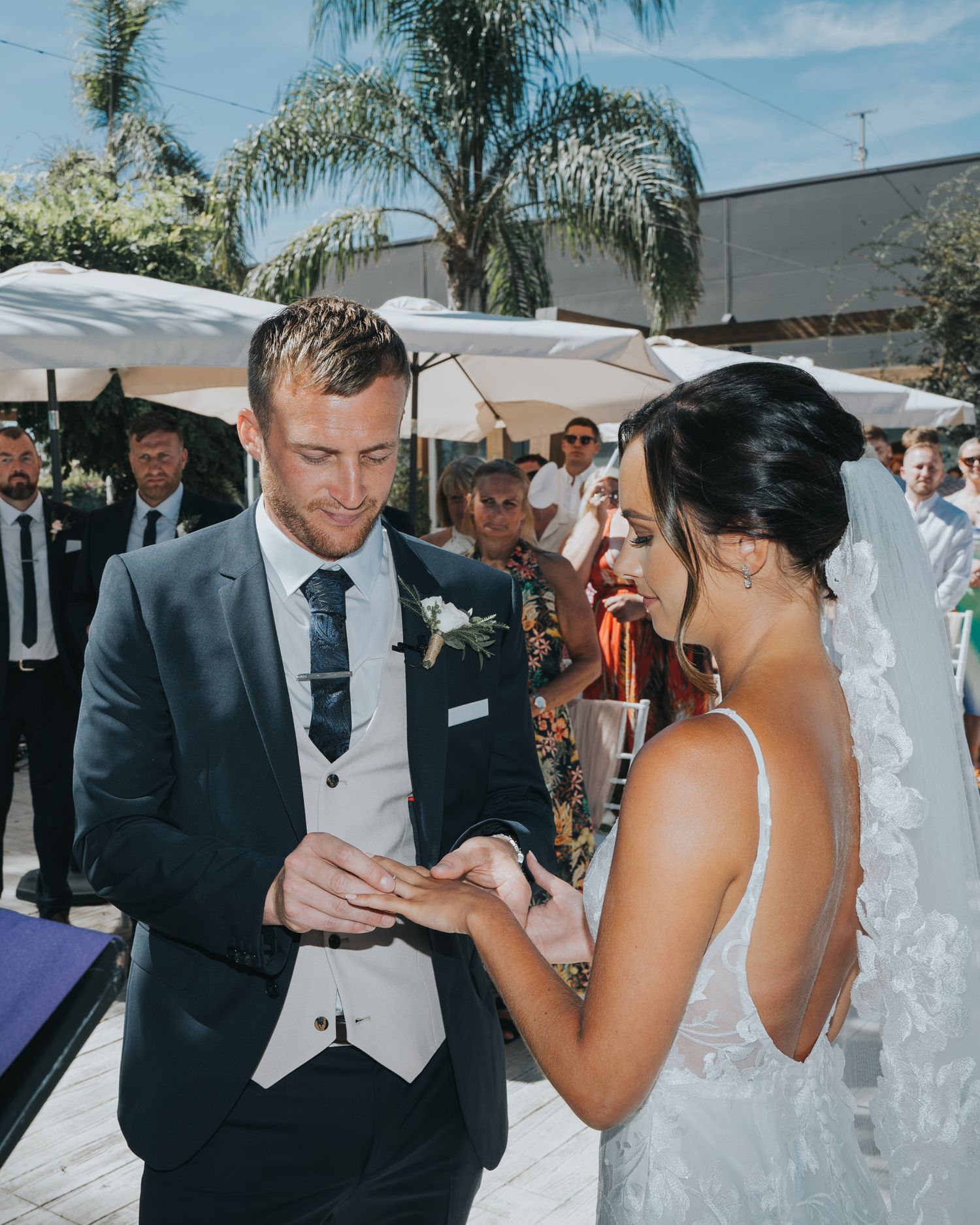 A couple exchanges rings during an outdoor wedding ceremony under white umbrellas and palm trees.