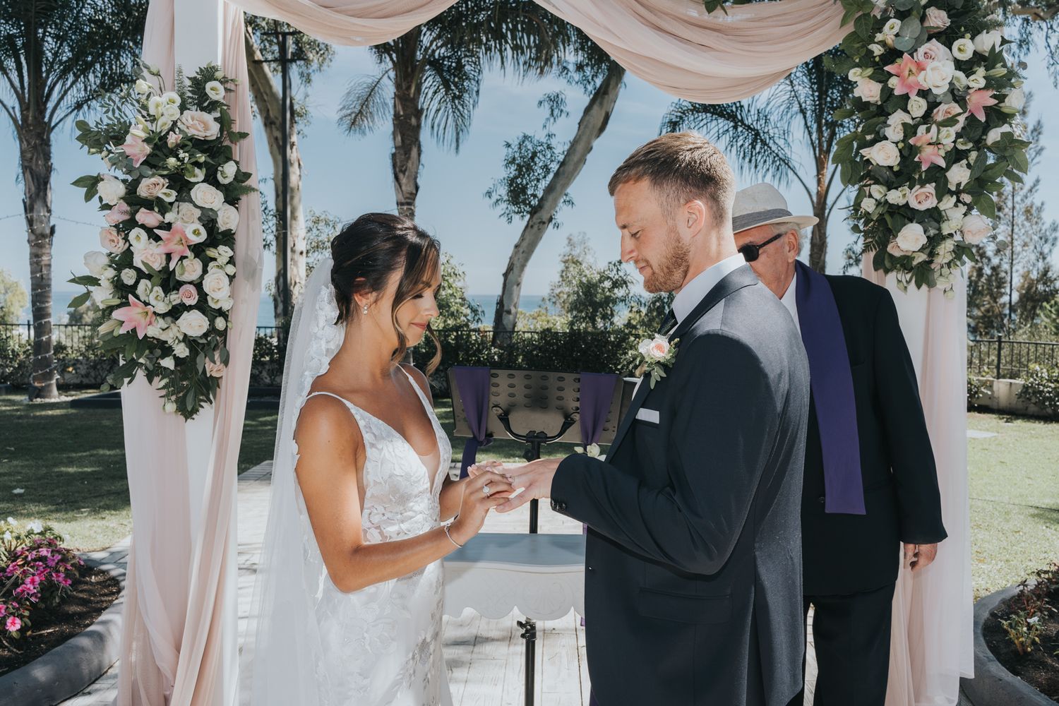Outdoor wedding ceremony under a floral arch with blush pink drapery.