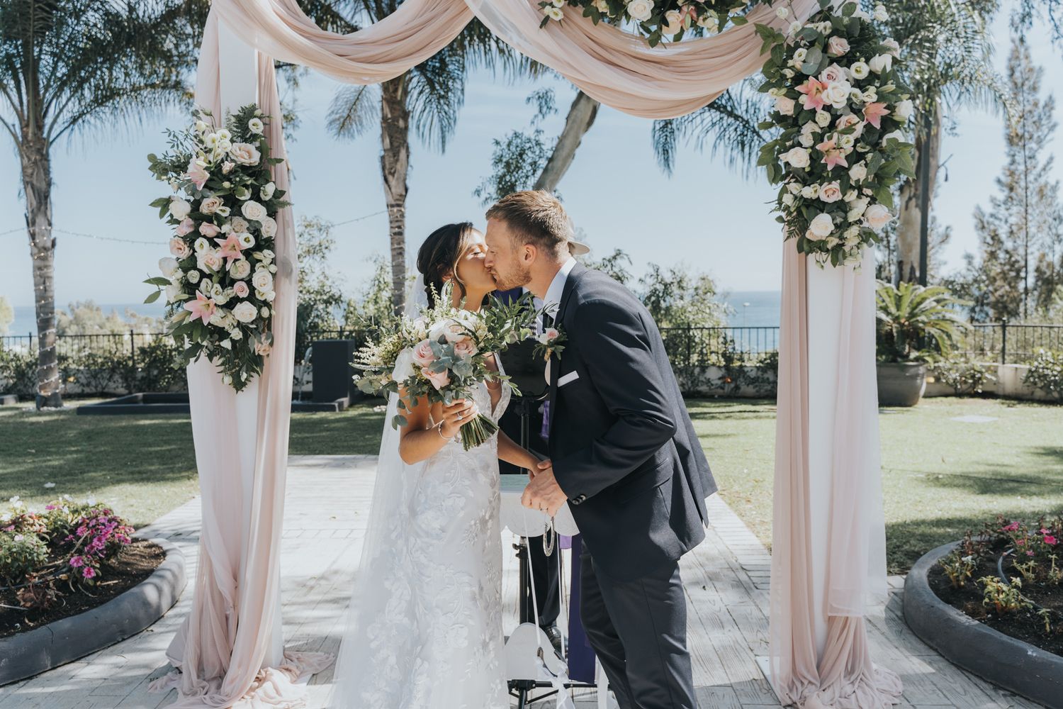 Wedding ceremony under a floral arch with blush drapery and garden landscape in background.