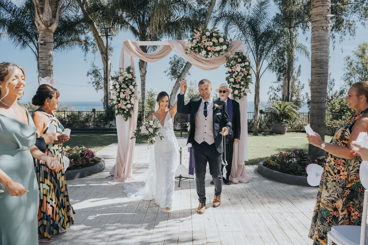 A wedding ceremony takes place under an elegant floral arch with palm trees and blue sky in the background.