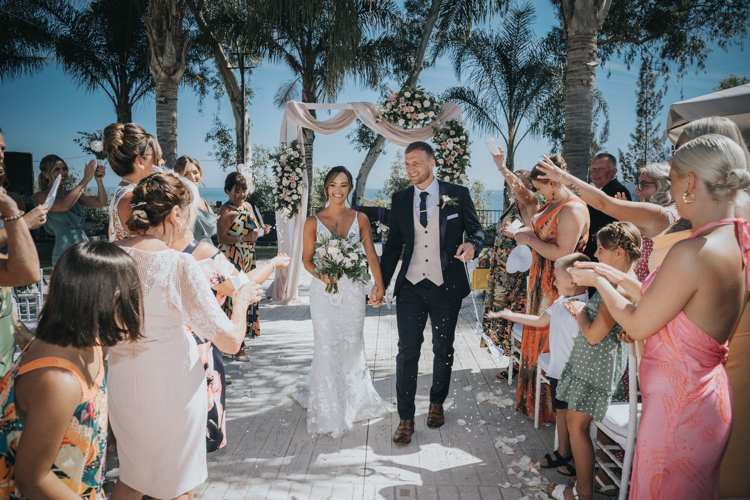 Wedding ceremony celebrations at an outdoor venue with guests tossing flower petals as newlyweds walk down palm-lined aisle.