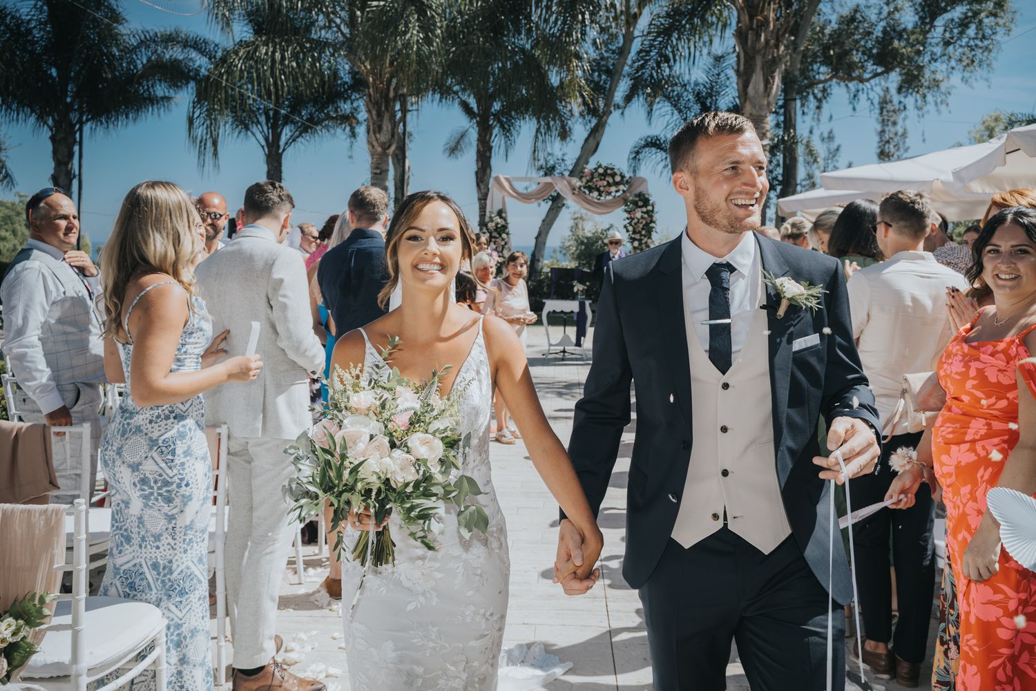A happy couple walks down the aisle during their tropical outdoor wedding ceremony with palm trees in the background.