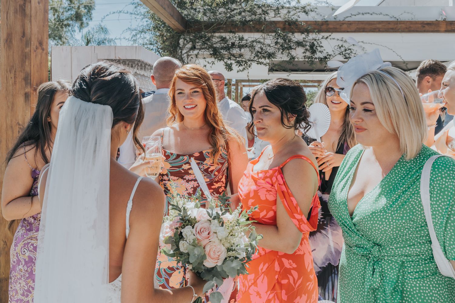 A bridal party laughs together while holding bouquets at an outdoor celebration.