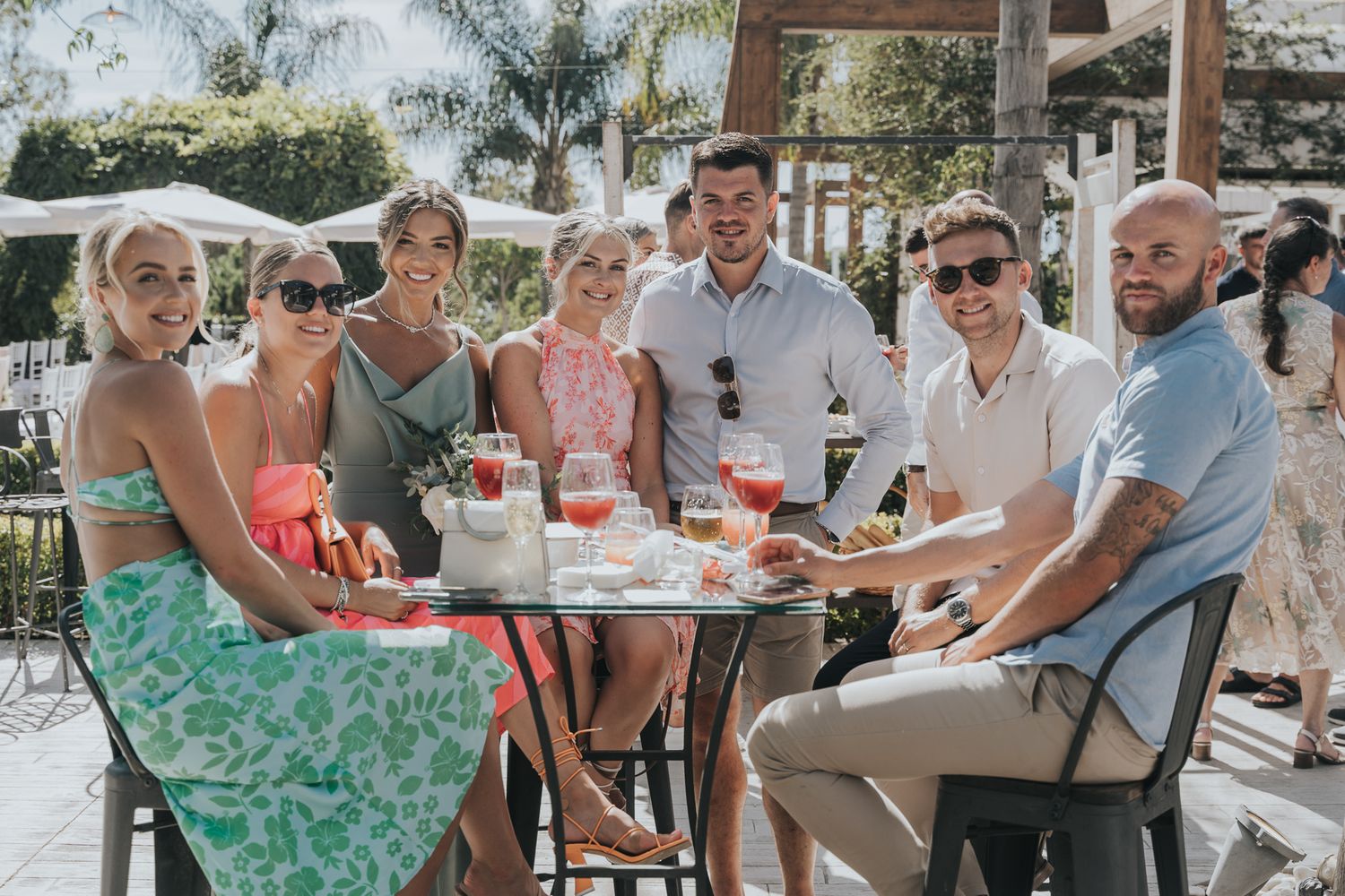 A group of guests enjoying drinks and conversation at an outdoor table during a sunny celebration.