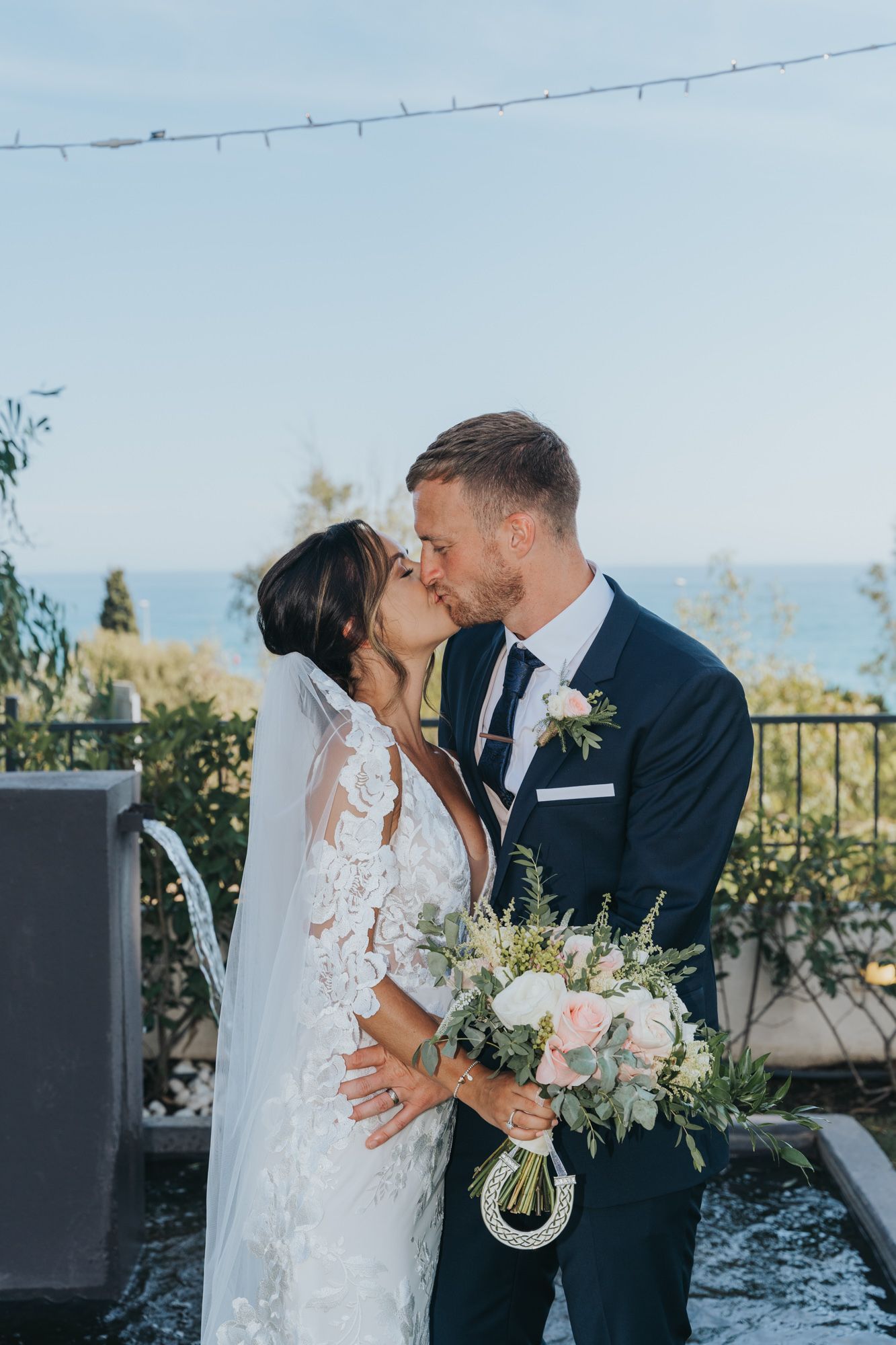A newlywed couple shares a romantic kiss on a terrace overlooking the ocean with a beautiful bouquet.
