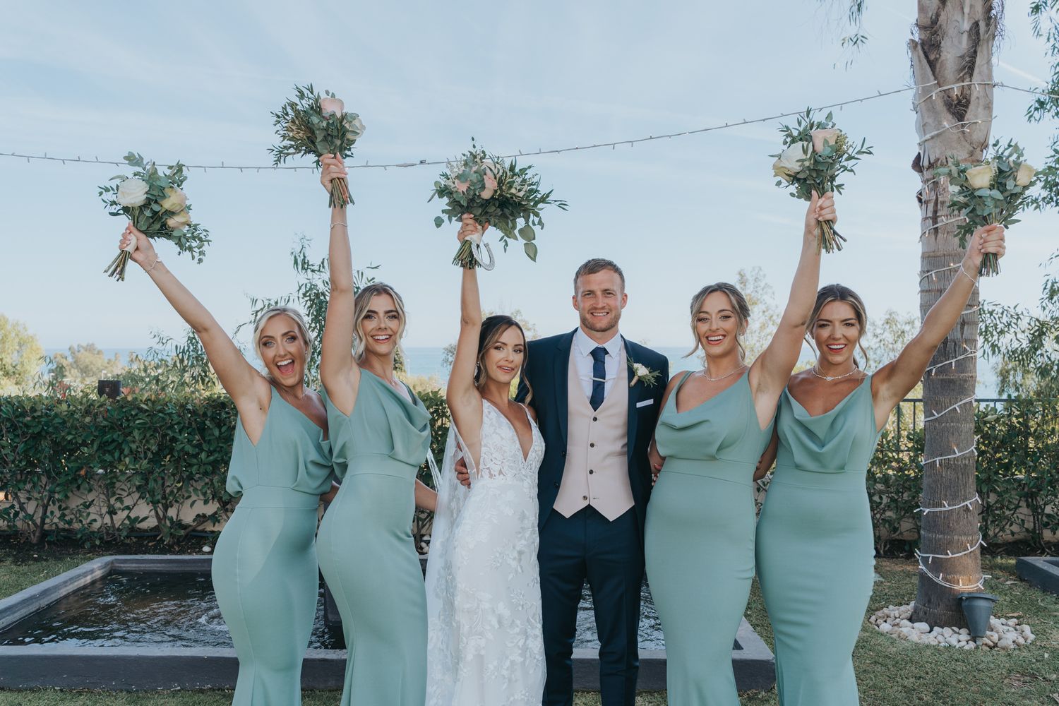 A wedding party poses together with bridesmaids in matching mint green dresses holding floral bouquets.