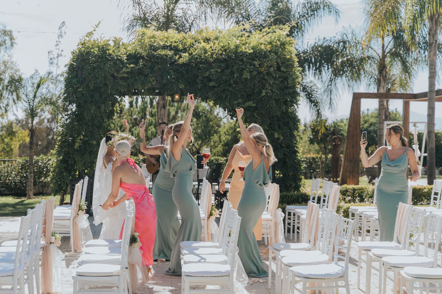 Bridesmaids in sage green dresses celebrating under a lush greenery arch at an outdoor wedding venue.