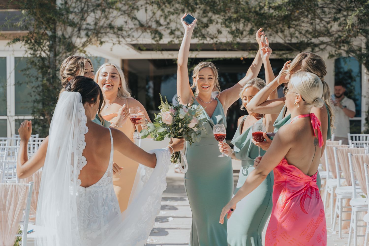 Bridesmaids in colorful dresses celebrate with dancing and laughter at an outdoor wedding venue.