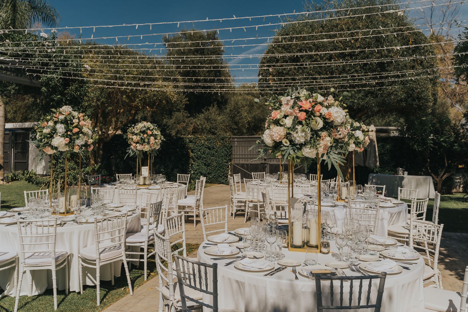 An elegant outdoor wedding reception setup with white tables, chairs and tall floral centerpieces under string lights.