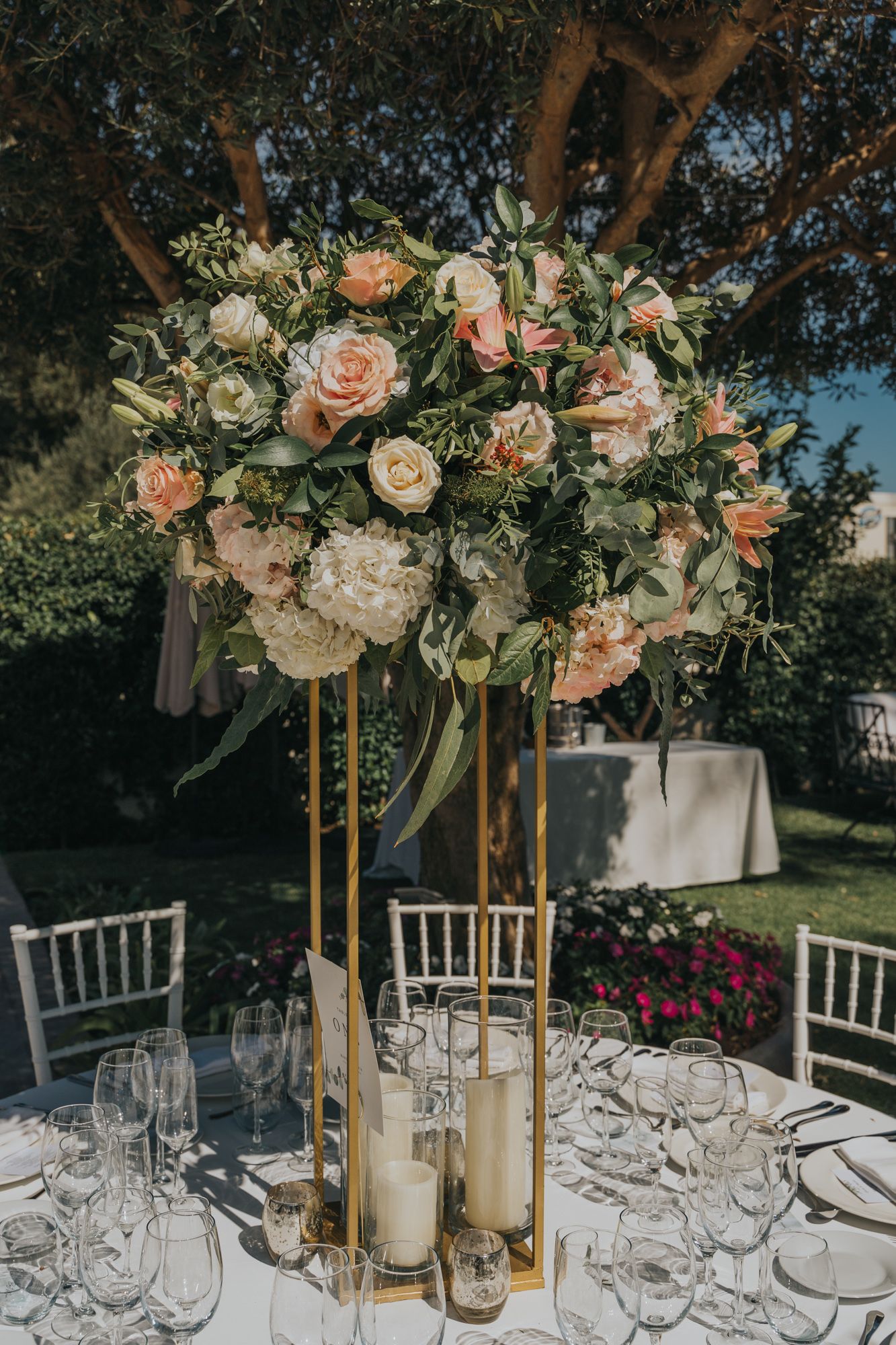 Elegant peach and white floral centerpiece on gold stand with table settings at outdoor wedding reception.