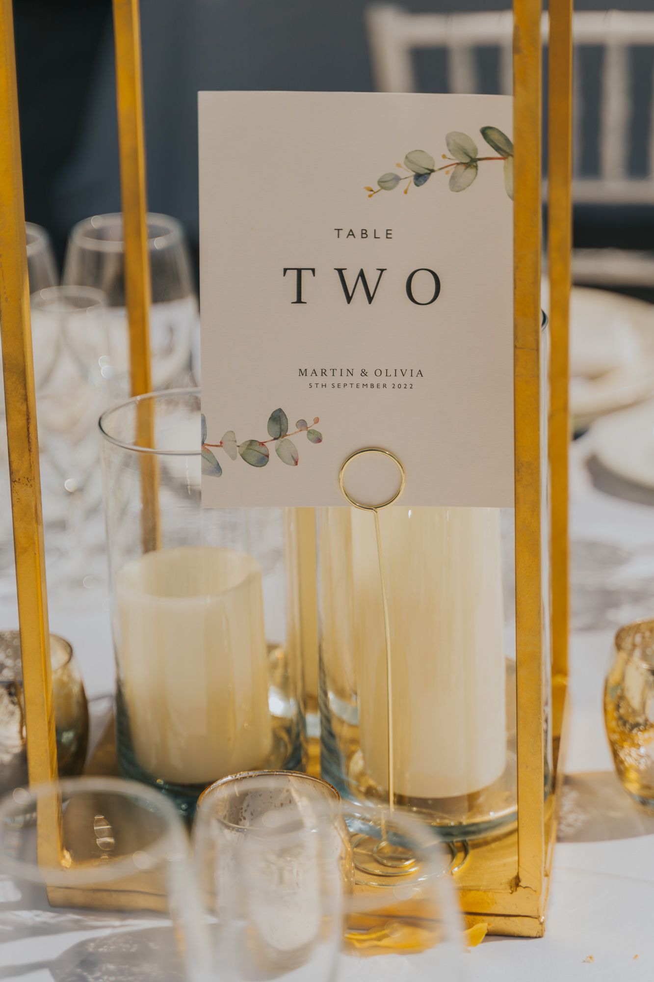 A minimalist table number card with eucalyptus design displayed between gold candlesticks at a wedding reception.