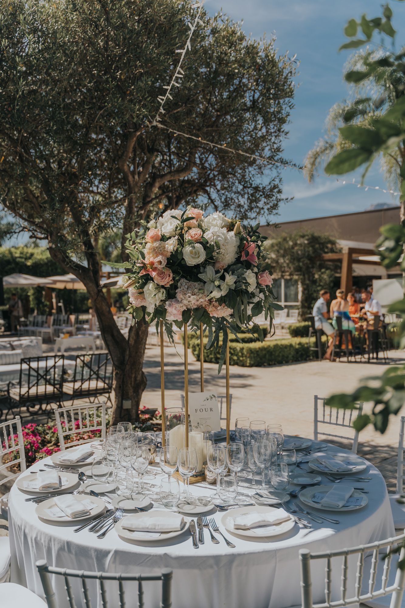 Elegant wedding table setting with tall floral centerpiece under an olive tree.