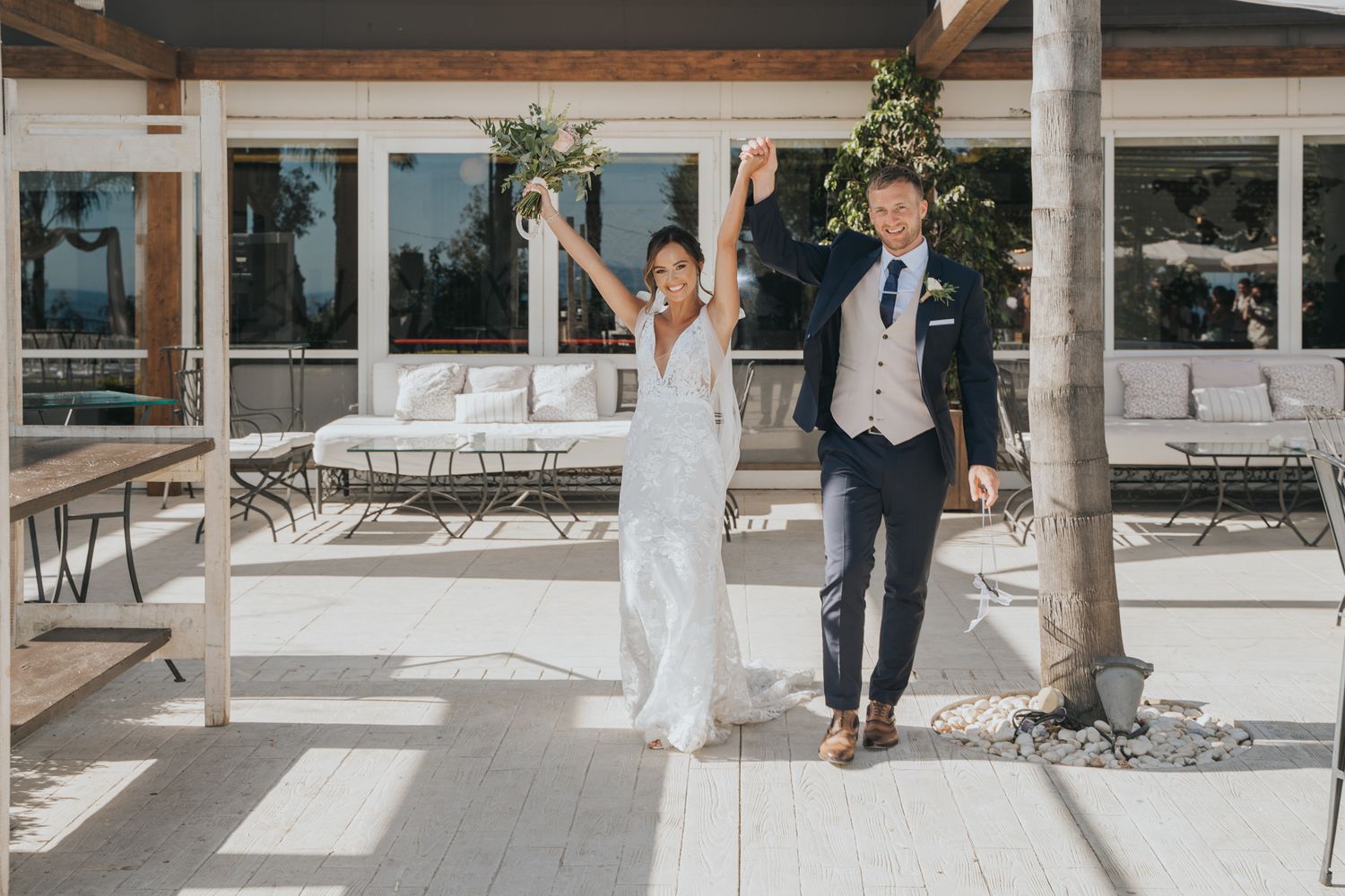 Wedding couple celebrates joyfully on a bright patio with white decor and outdoor seating.