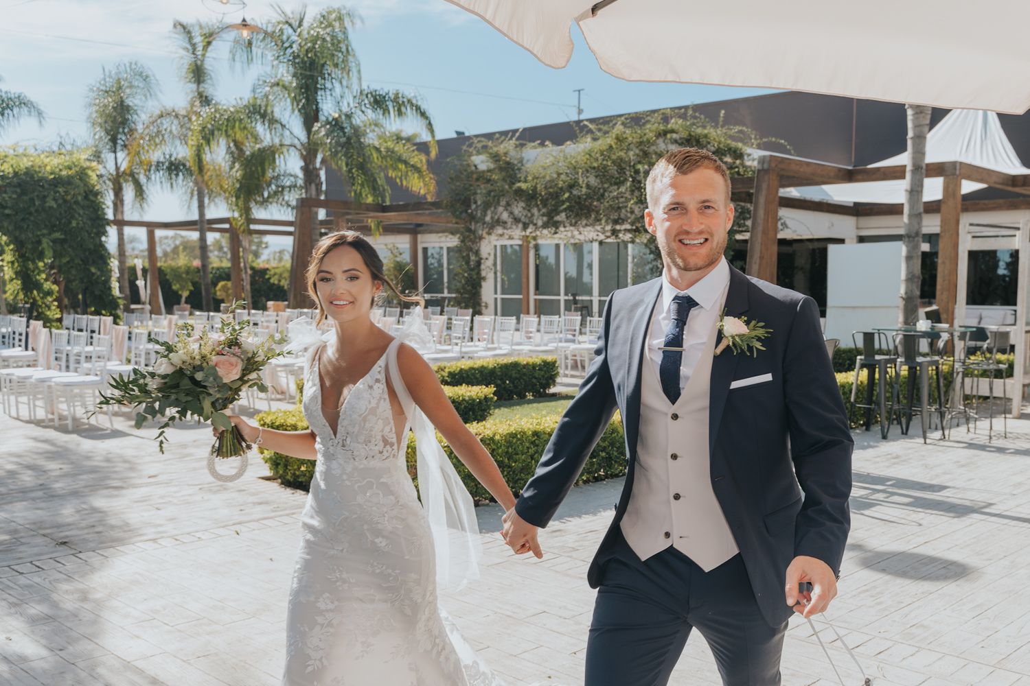 A newlywed couple walks hand in hand through their outdoor venue with palm trees in the background.