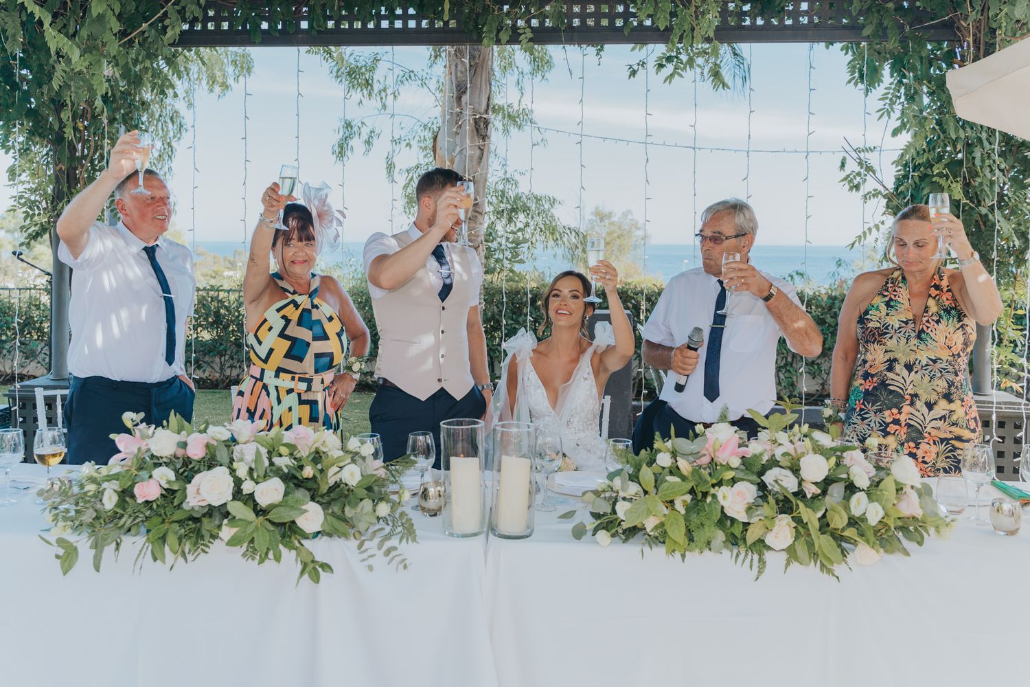 Wedding party celebrates at a beautifully decorated head table with floral arrangements and greenery.