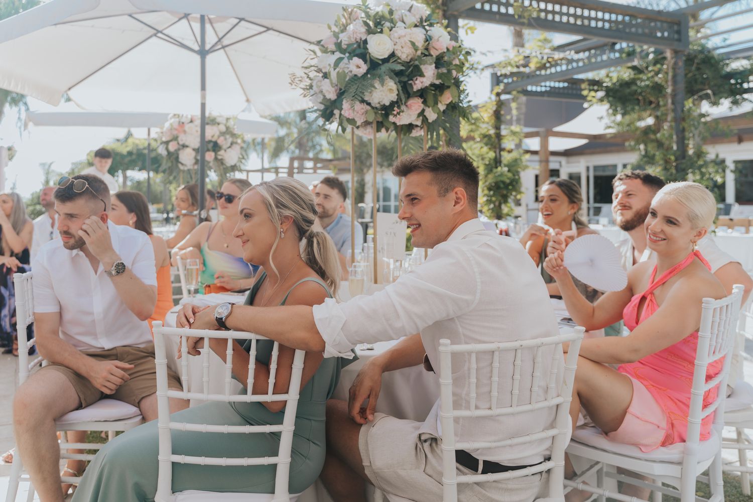 Outdoor wedding reception with guests seated at tables under white umbrellas and floral arrangements.