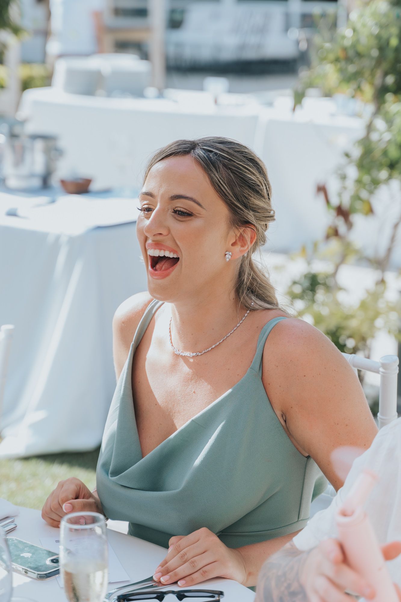 Guest in sage green dress laughing joyfully at outdoor wedding reception.