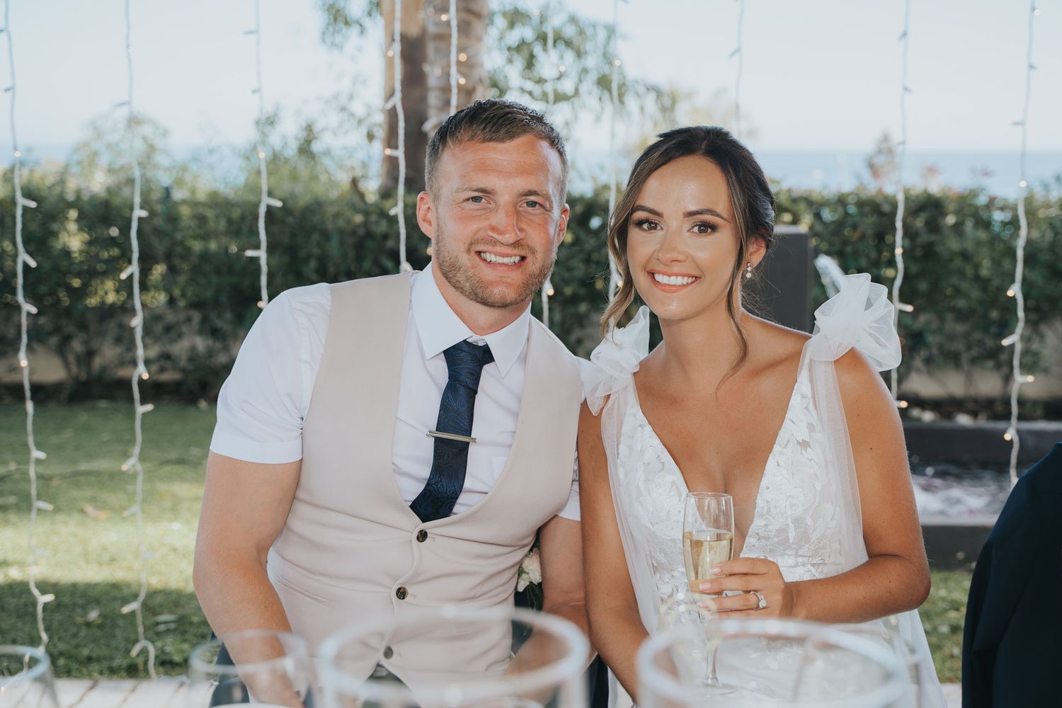 A couple in wedding attire sit together and smile while holding champagne glasses at an outdoor reception.