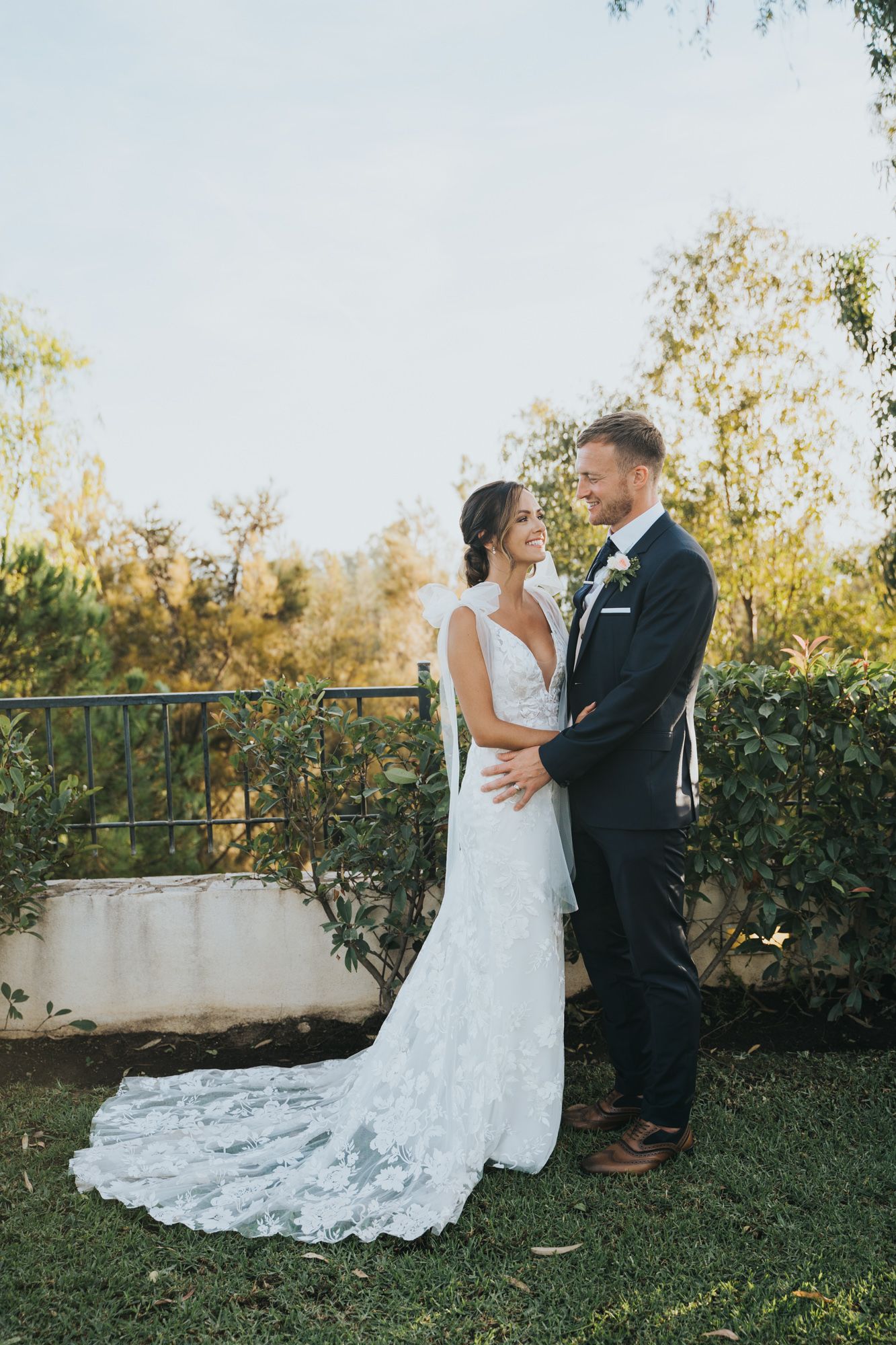 Elegant wedding couple embraces on grassy terrace with bride wearing lace train dress.