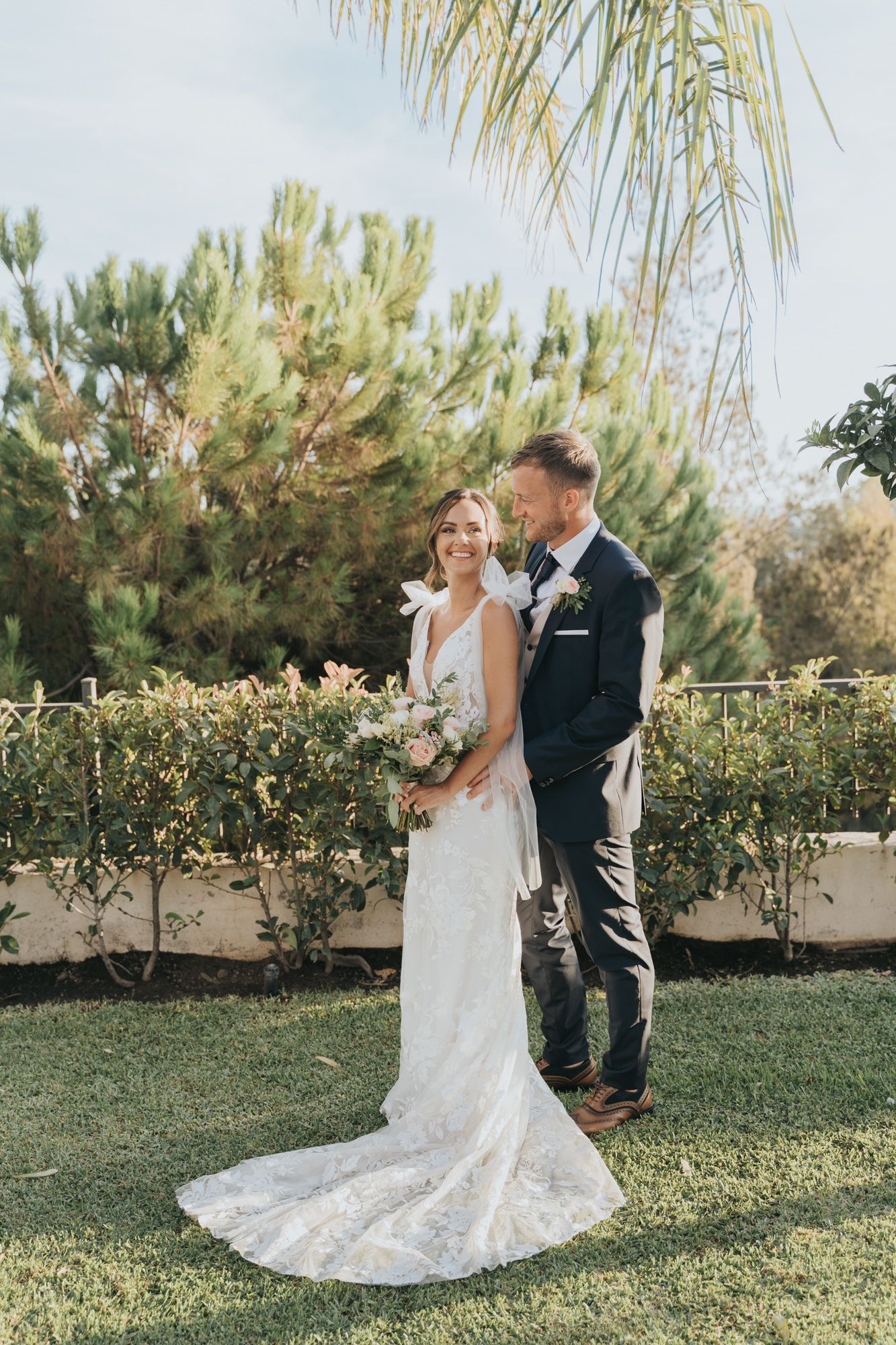 A couple poses together in formal wedding attire against a backdrop of greenery.