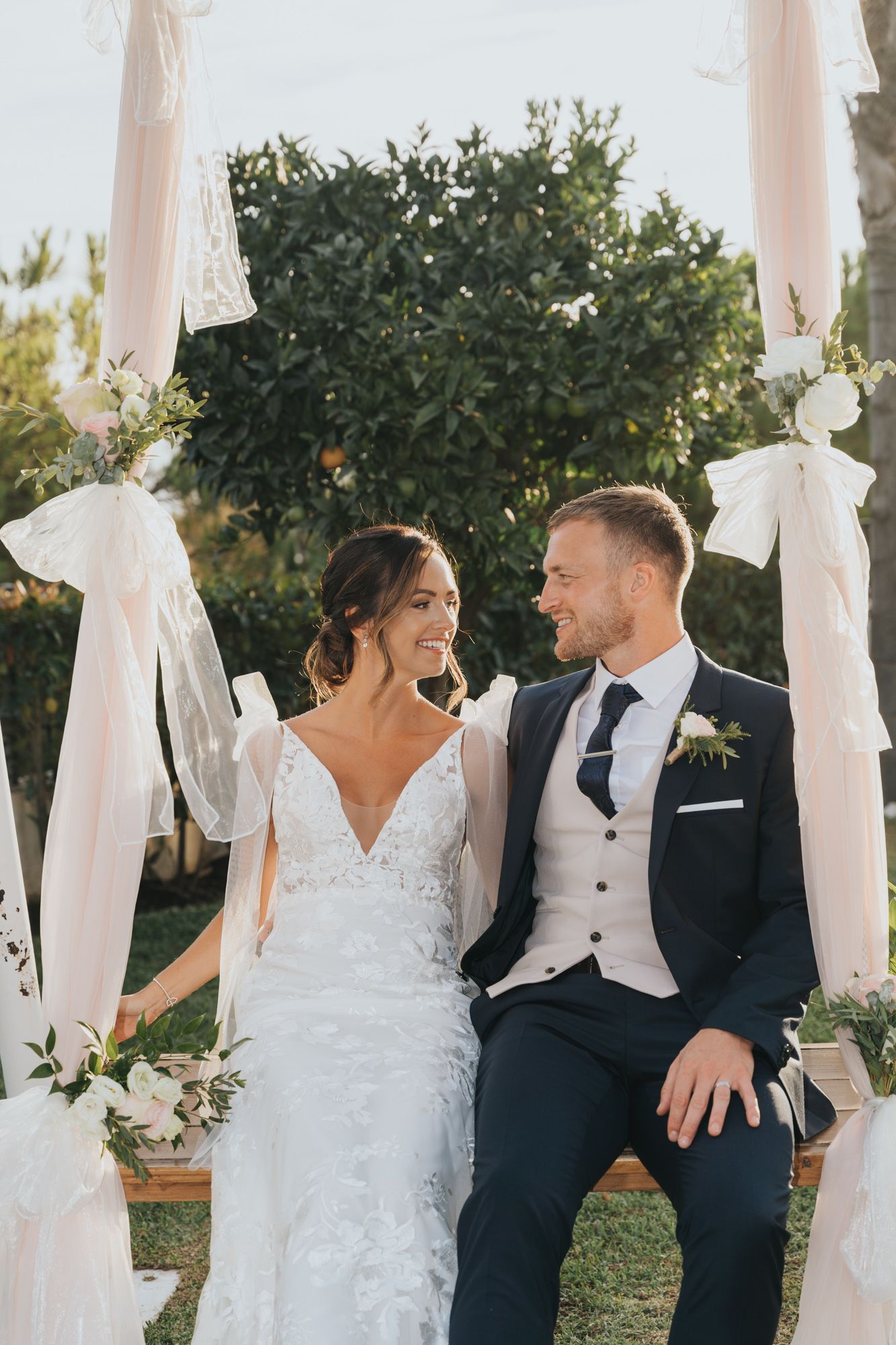 Couple sitting together on decorated wedding bench with white drapes and floral arrangements.