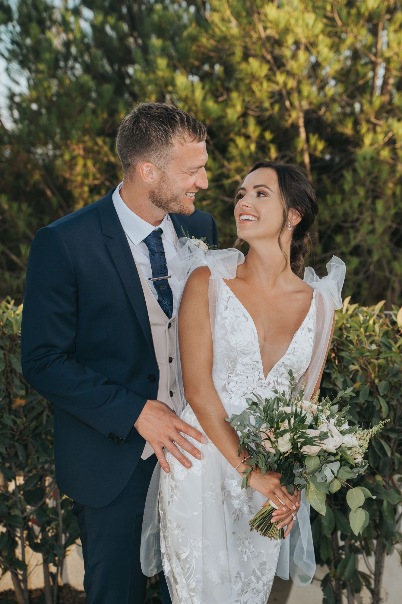 A romantic wedding photo shows a couple in formal attire sharing a loving moment against a green outdoor backdrop.